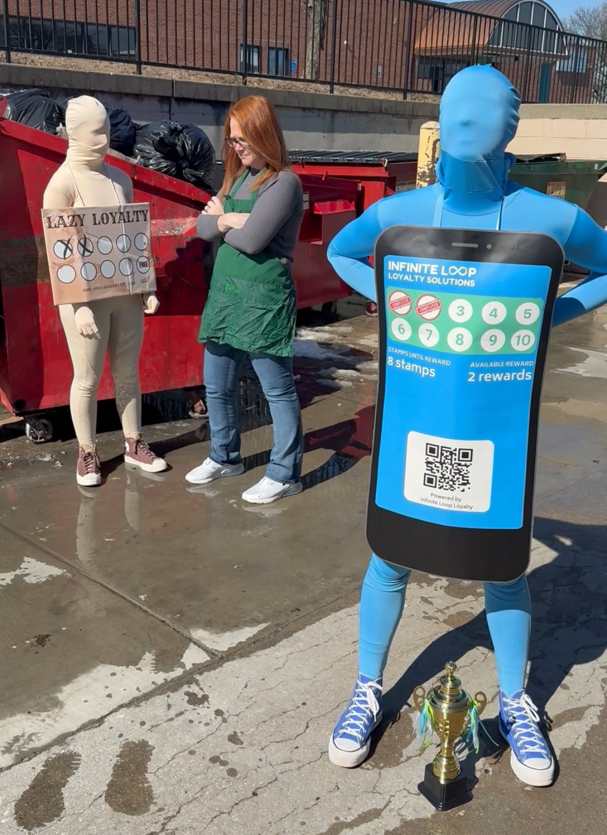 Costumed characters representing a lazy paper punch card and a digital loyalty app stand outside a dumpster, with the digital loyalty mascot holding a trophy labeled Infinite Loop Loyalty Solutions.