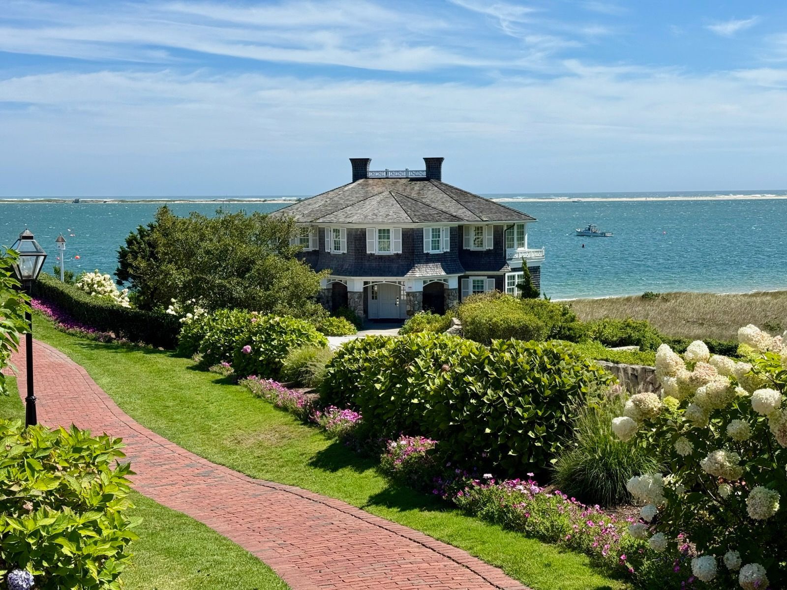 A red brick path leads toward a grey, shingled house on the coast, surrounded by lush green shrubs and white flowers.