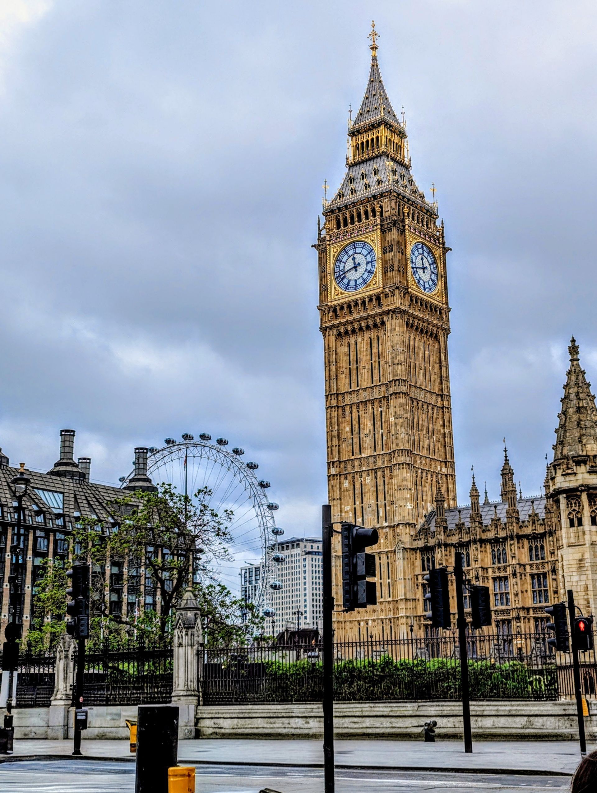 Big Ben clock tower and the Houses of Parliament under a cloudy sky in London, with the London Eye visible in the distance.