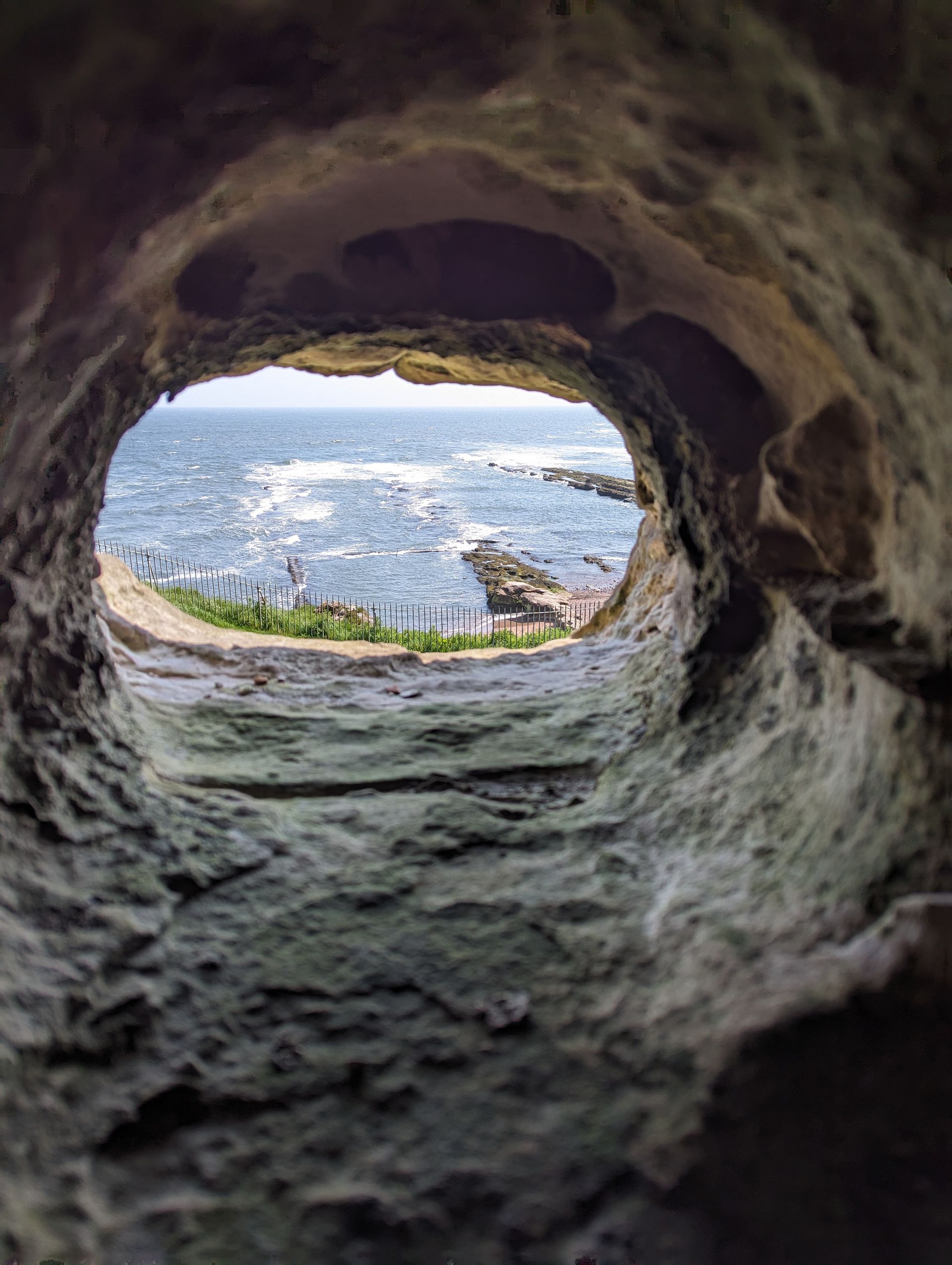 View of a blue ocean and coastline through a rough, circular stone opening in a rock formation.