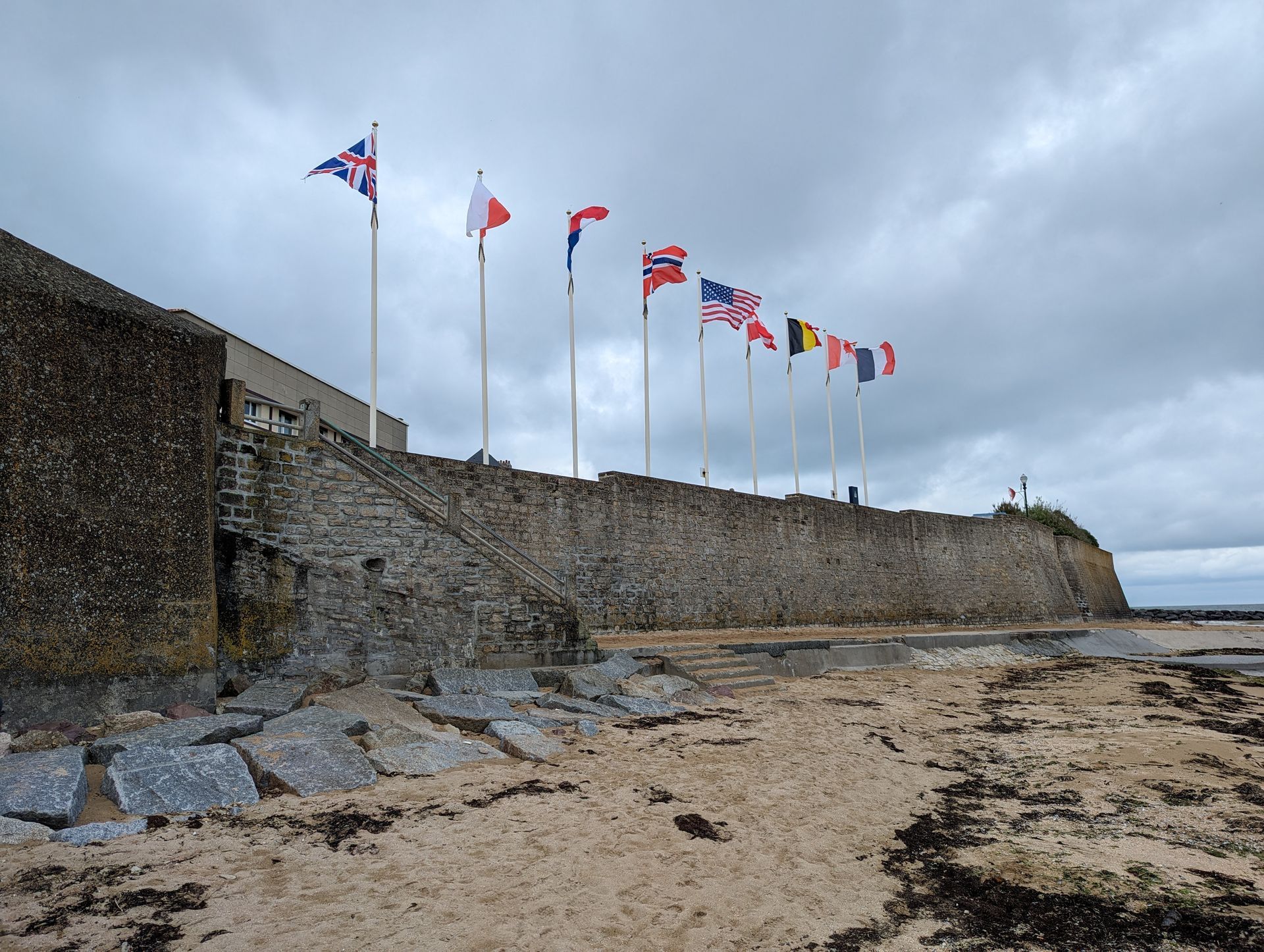 A stone sea wall topped with a row of various national flags flying under a cloudy sky.