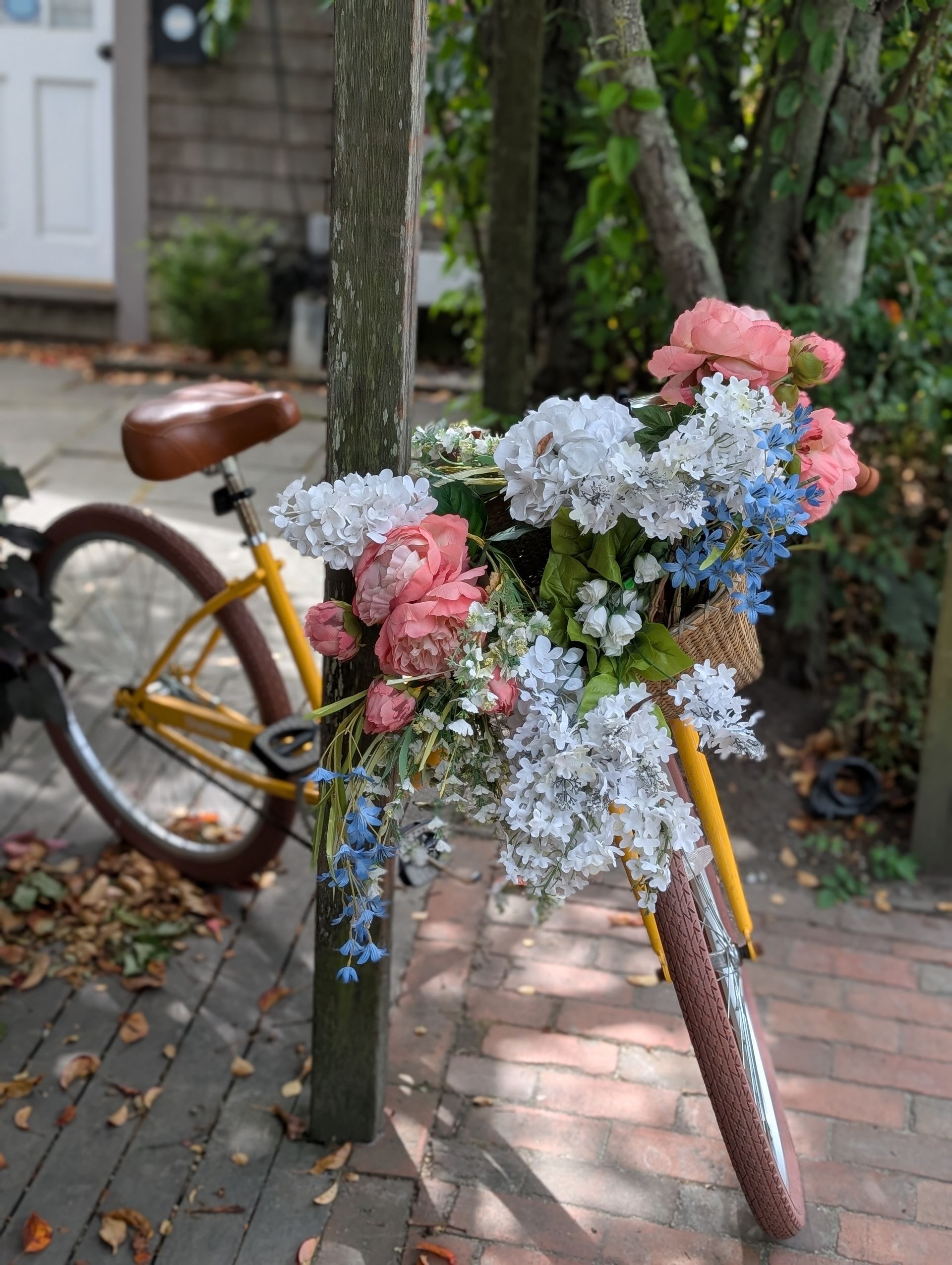 A yellow bicycle with a large basket of white, pink, and blue flowers leaning against a post on a brick path.