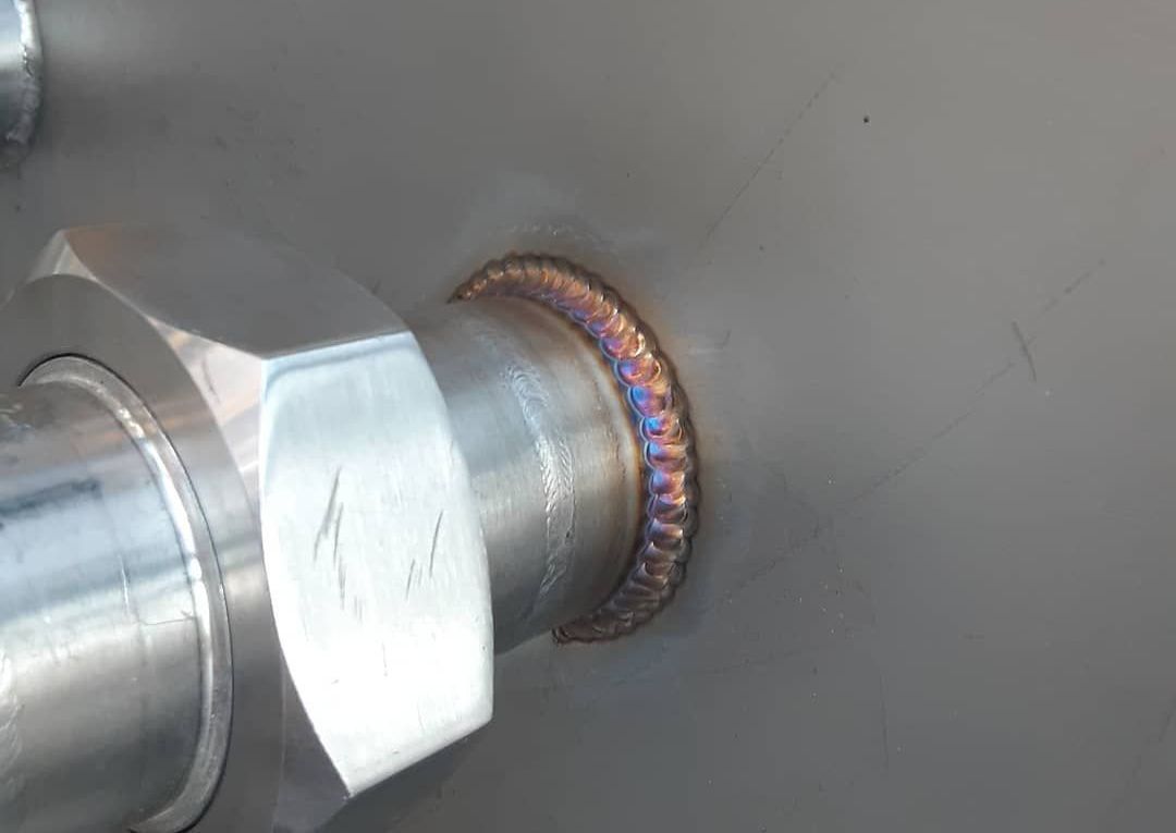 A stainless steel nut welded to a metal surface. The weld is colored with heat tints of blue and purple.