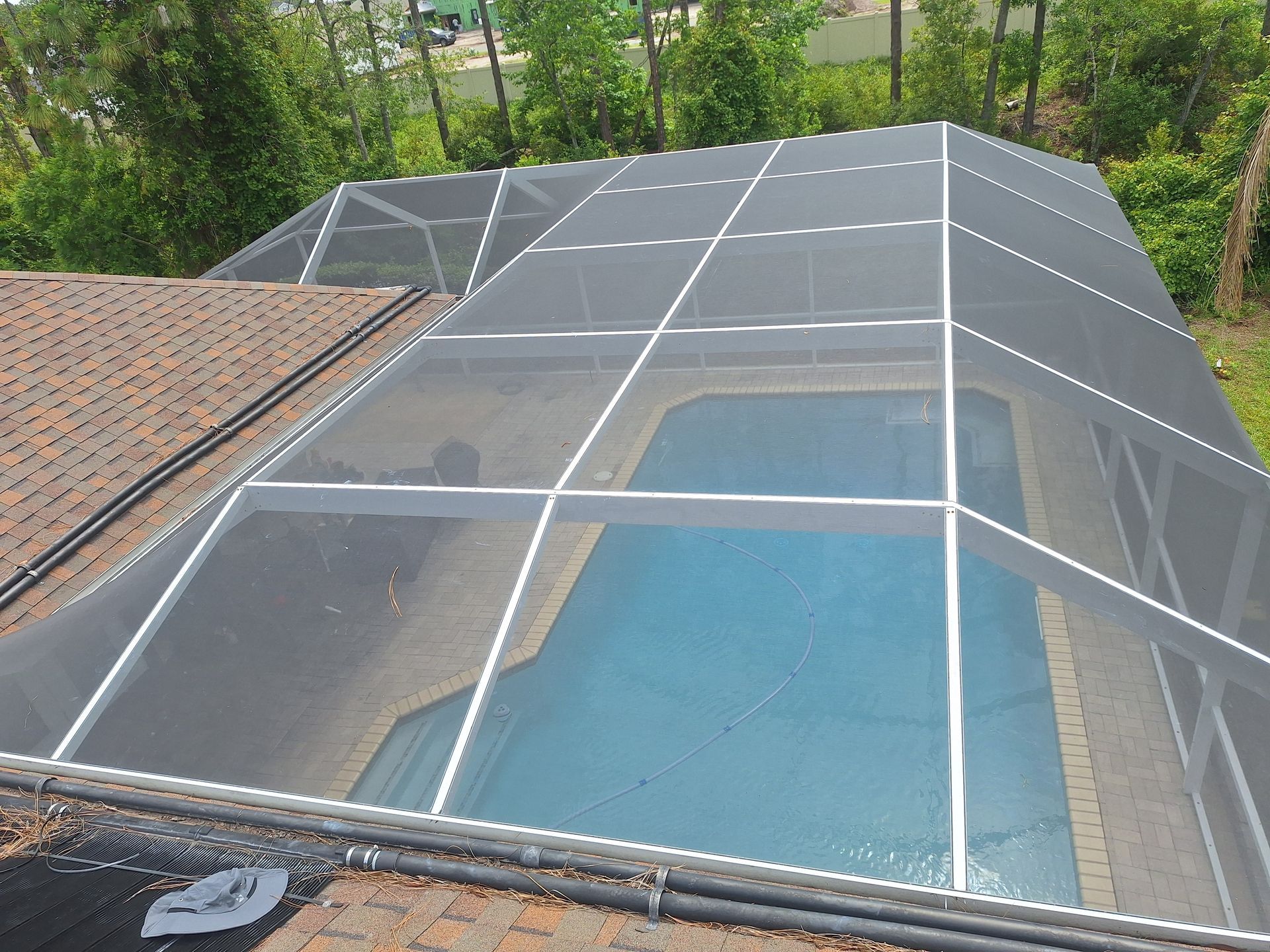 Pool enclosure with white framing and mesh roof over a blue pool, surrounded by a brick patio, viewed from above.