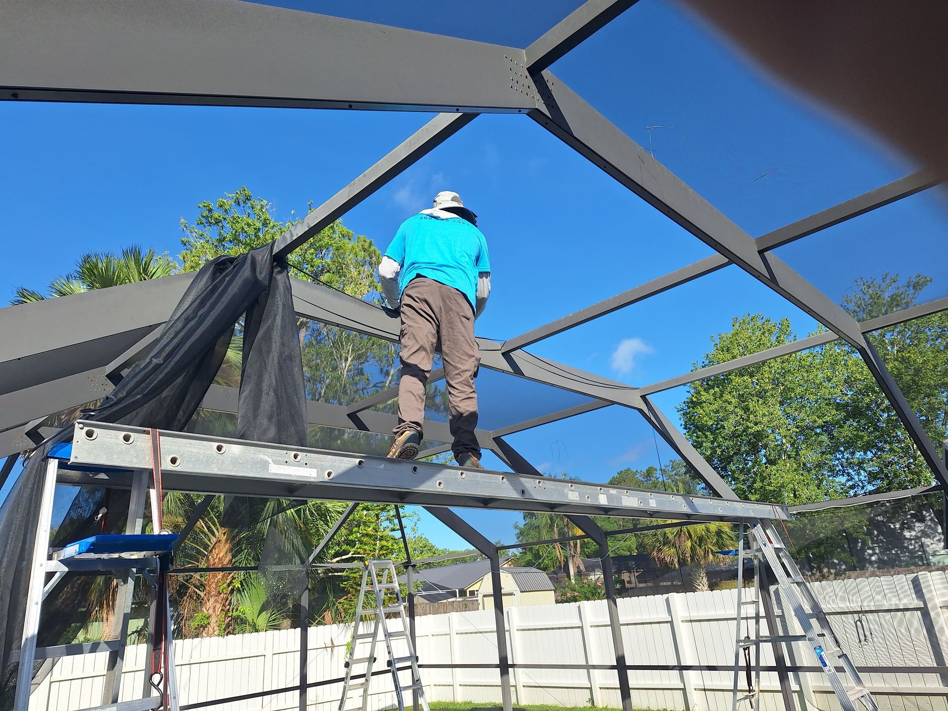Man on ladder repairing screen enclosure, sunny day.