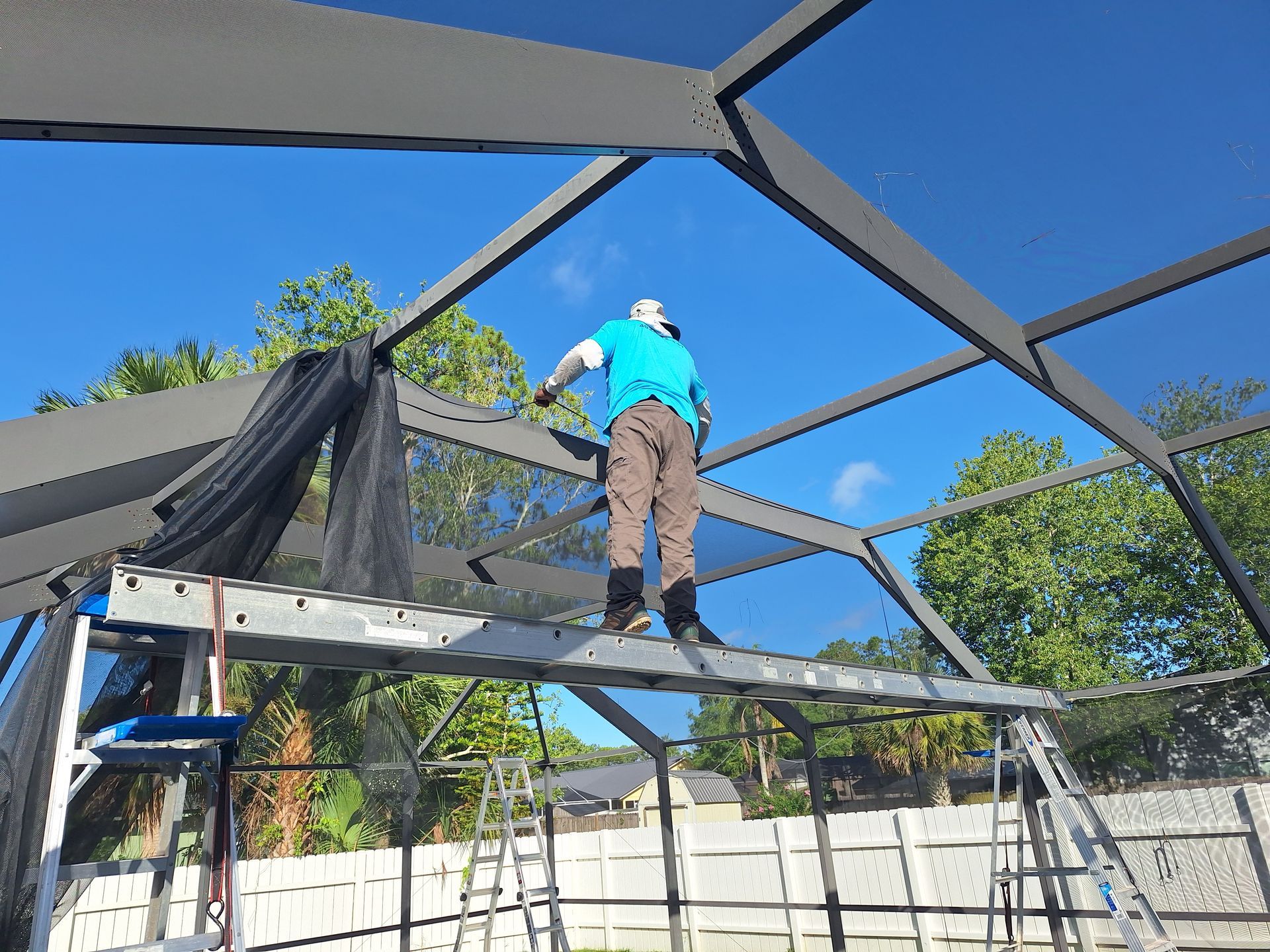 A worker on a scaffold repairs a screen enclosure roof against a blue sky.