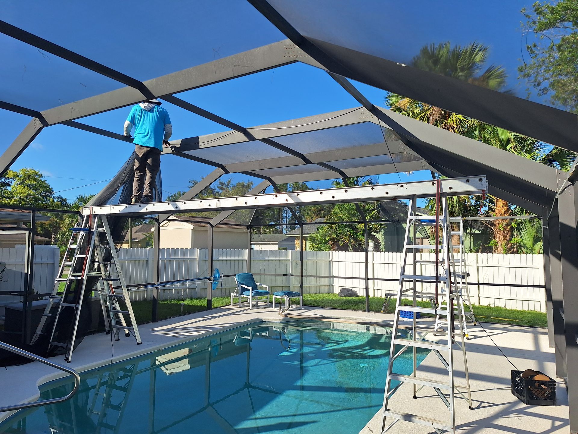 Man replacing pool screen under a clear blue sky, standing on a ladder.