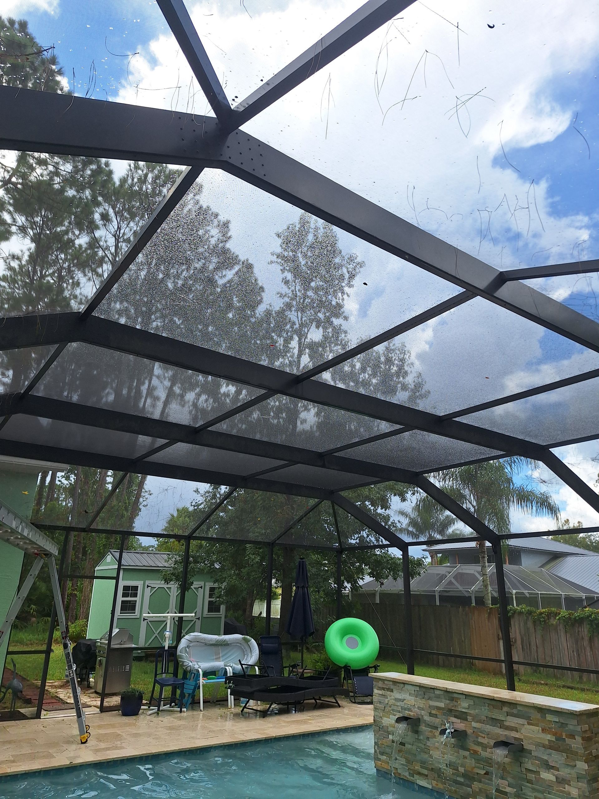 Screened-in patio over a pool. Black metal frame with netting. Blue water, green lawn, and trees visible.