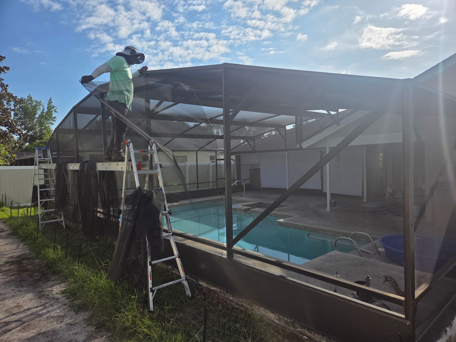Person on a ladder repairing a screen enclosure over a swimming pool. Sunny, blue sky.