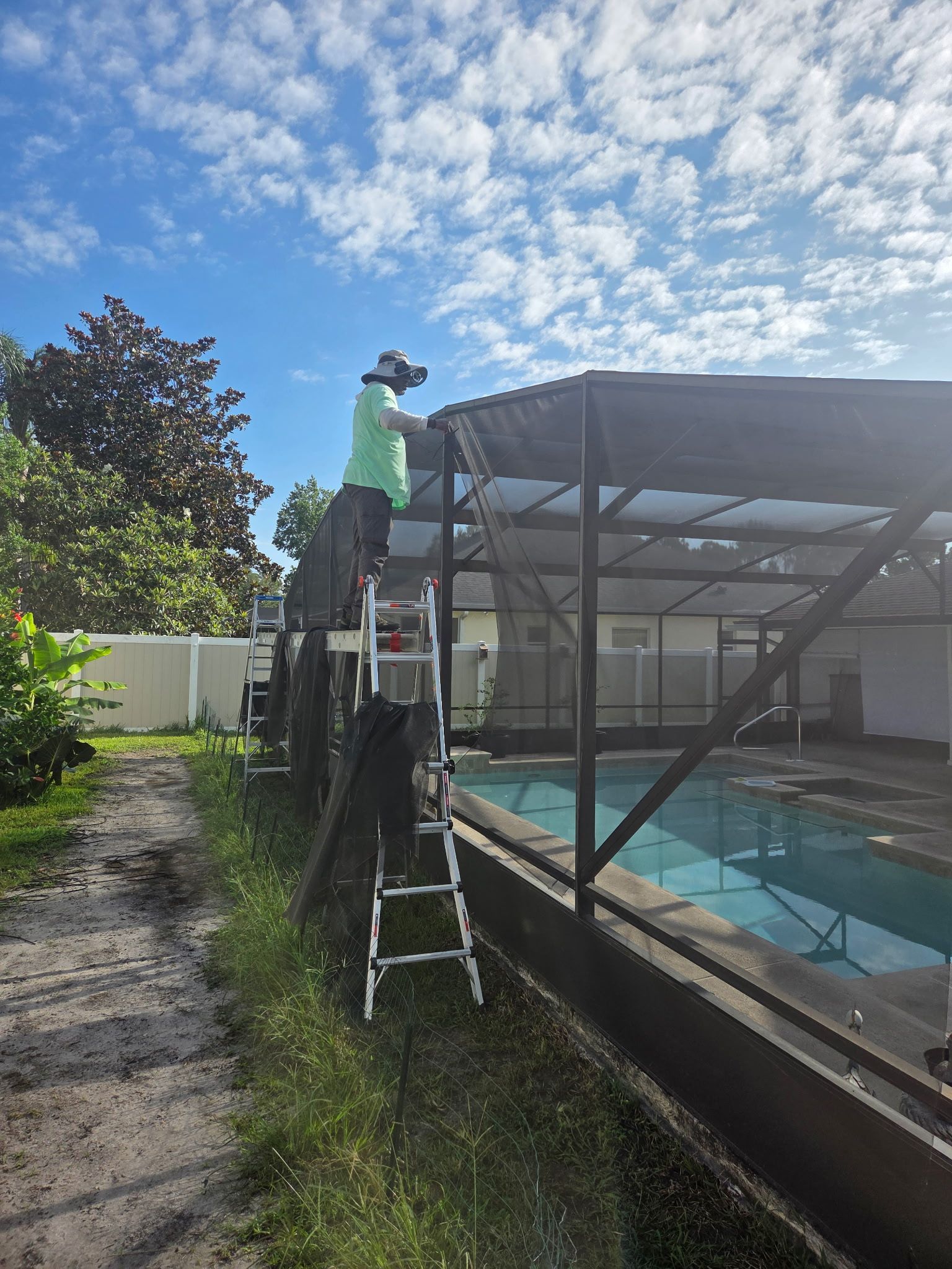 Person on ladder repairs pool screen enclosure under a blue sky.