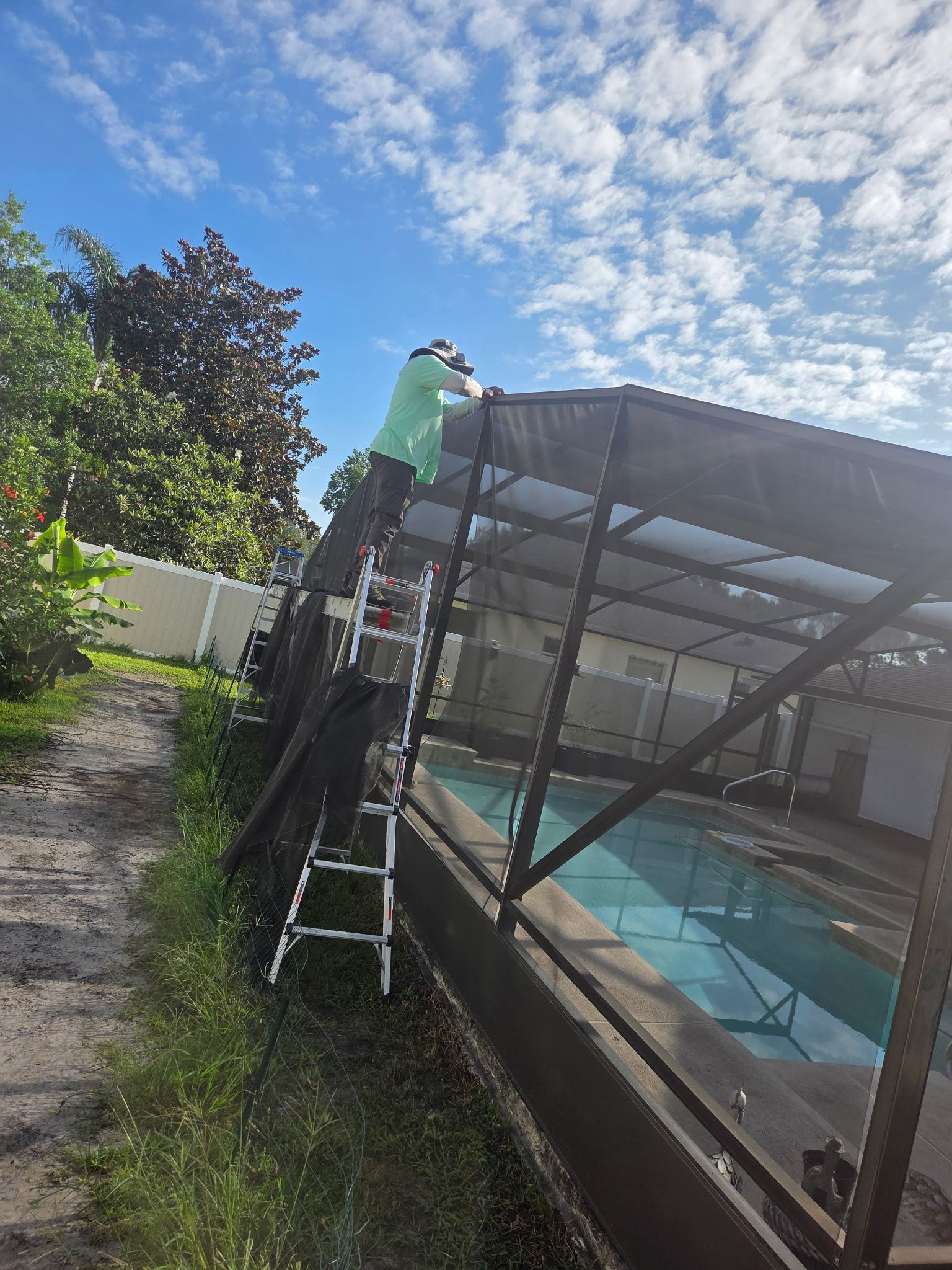 Person on ladder repairing screen enclosure over a swimming pool. Green clothing, sunny day.