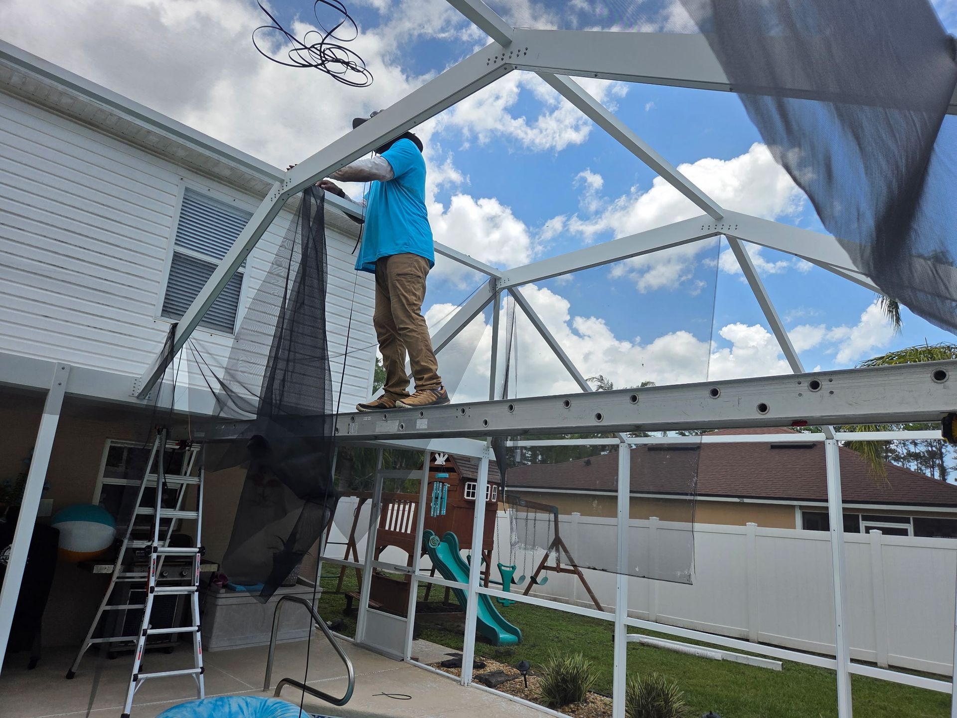 Man on a scaffold repairing screening on an enclosed patio; house in background.