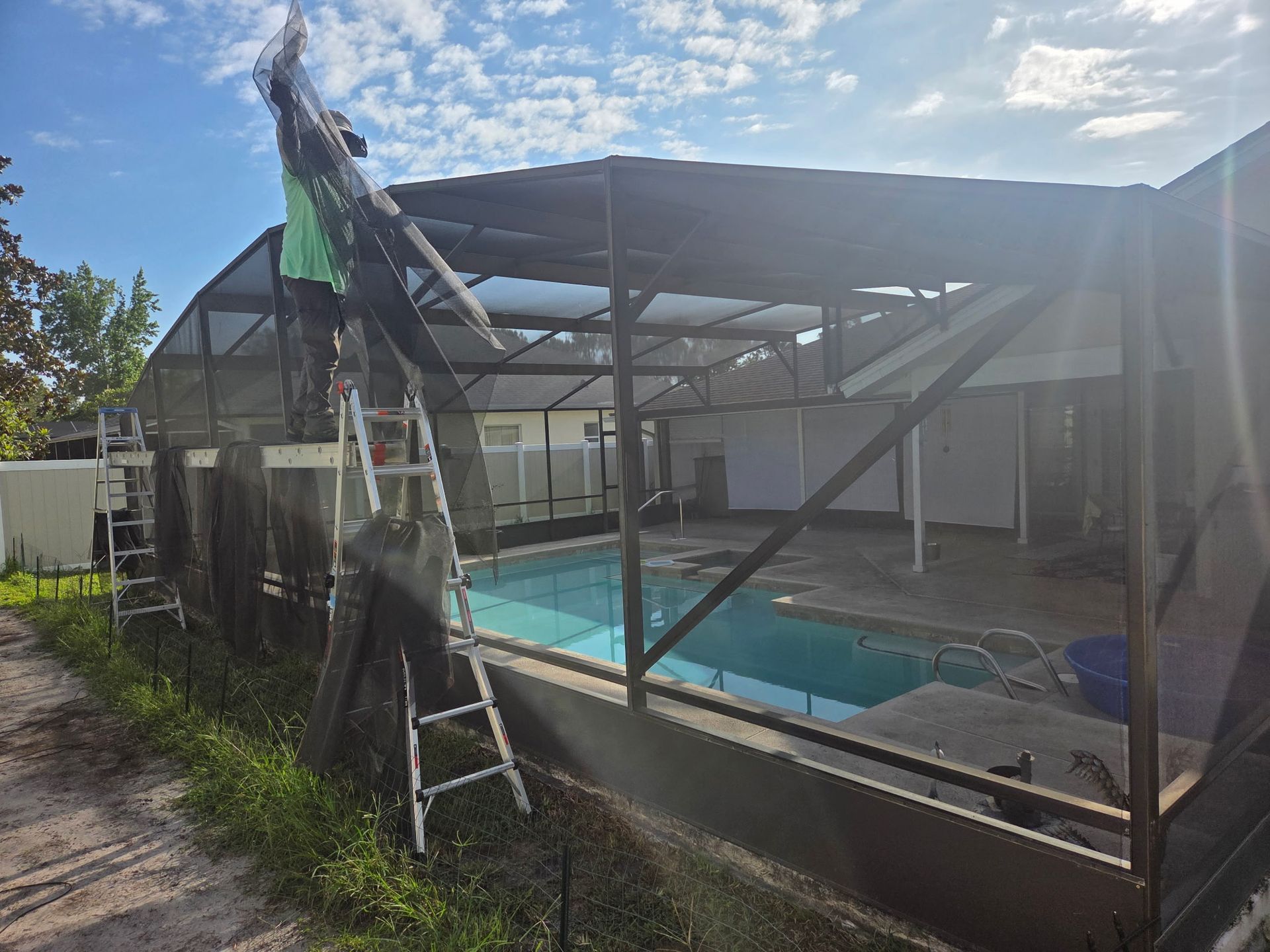 Person on ladder repairs pool enclosure screen. Pool visible, sunny outdoor setting.