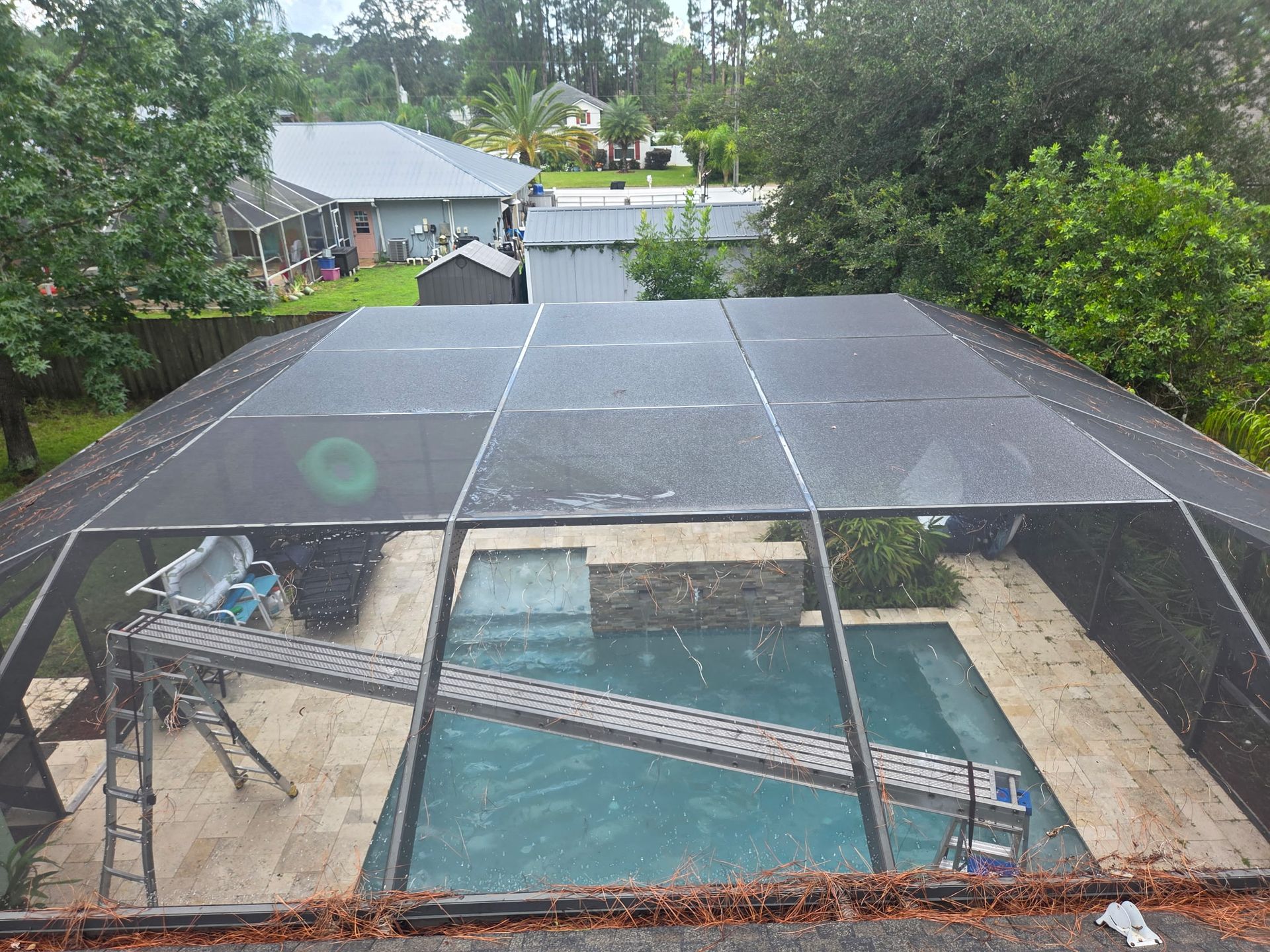 Pool with screen enclosure. Two ladders and scaffolding inside. Green trees and houses in the background.