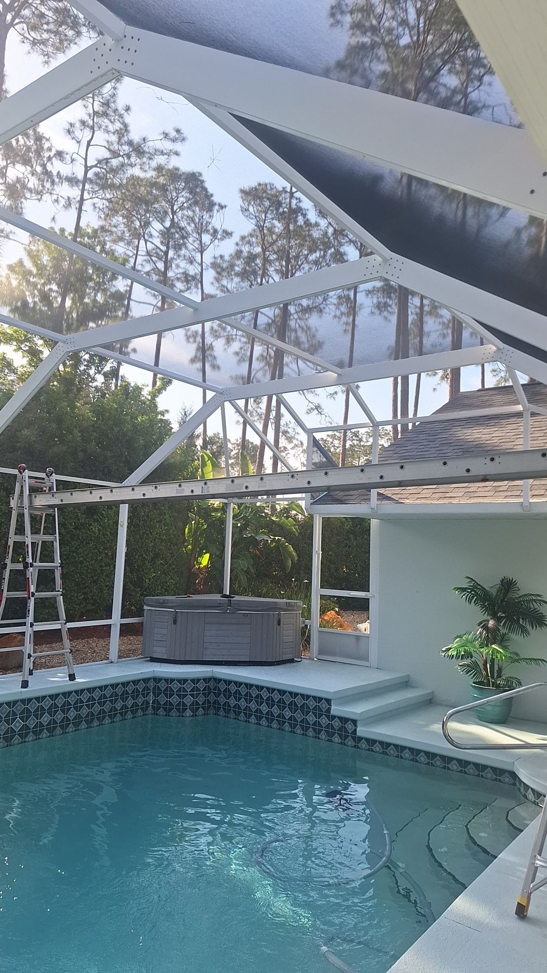 Pool enclosure with a swimming pool, hot tub, and ladder. Trees and sky visible through the screen.