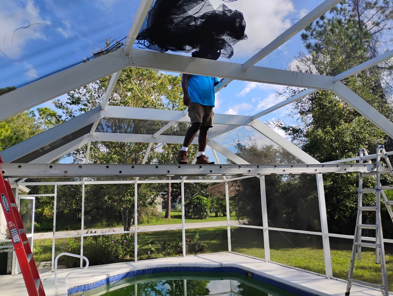 Person on a pool enclosure roof removing debris. White frame, clear panels, green pool, blue sky.