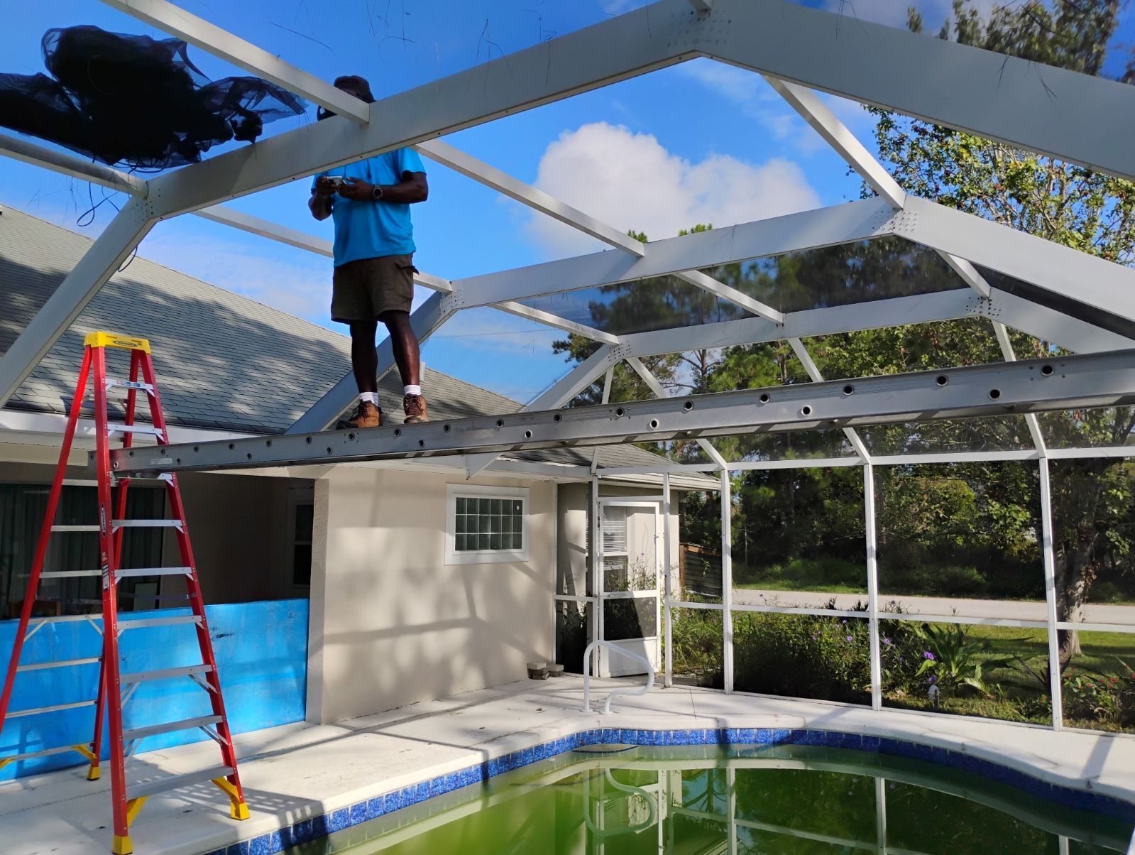 Person repairing screen enclosure over a pool. Ladder, blue sky, and green water visible.