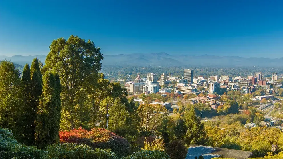 A view of a city from a hill with trees in the foreground and mountains in the background.