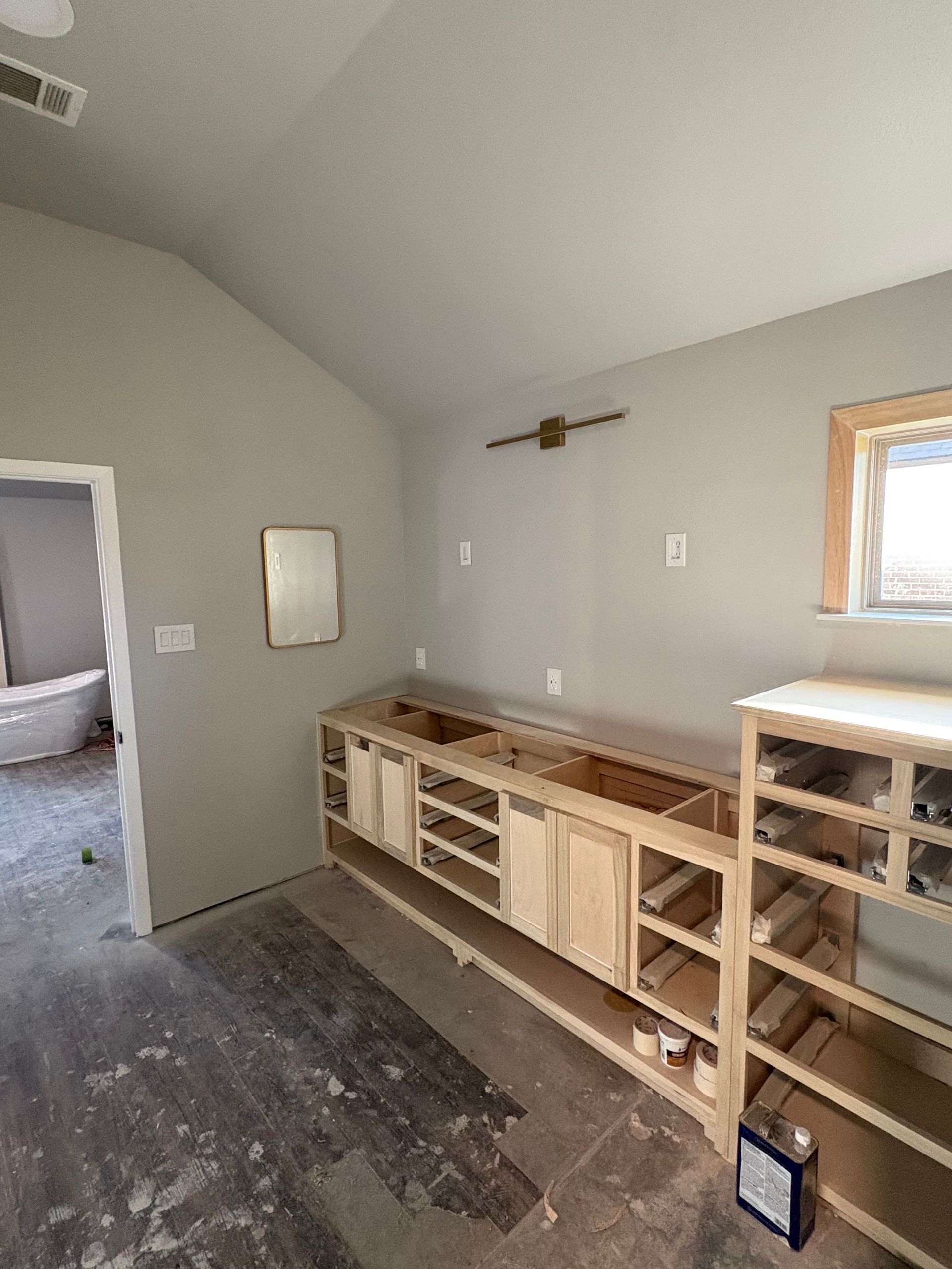 Unfinished bathroom: Light wood cabinets, gray walls, doorway to a toilet, window on right.