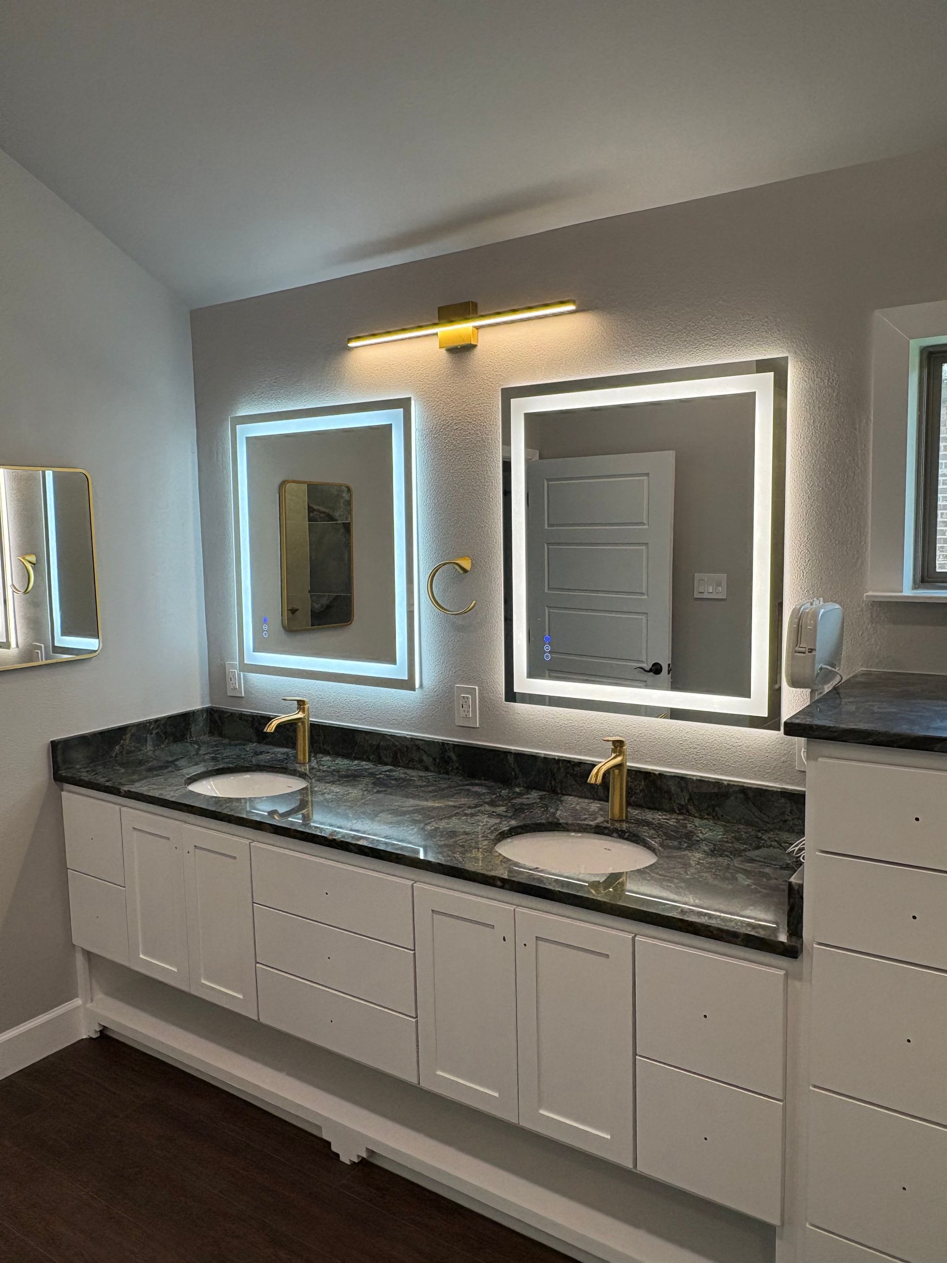 Modern bathroom with white cabinets, granite countertop, gold fixtures, and lit mirrors.