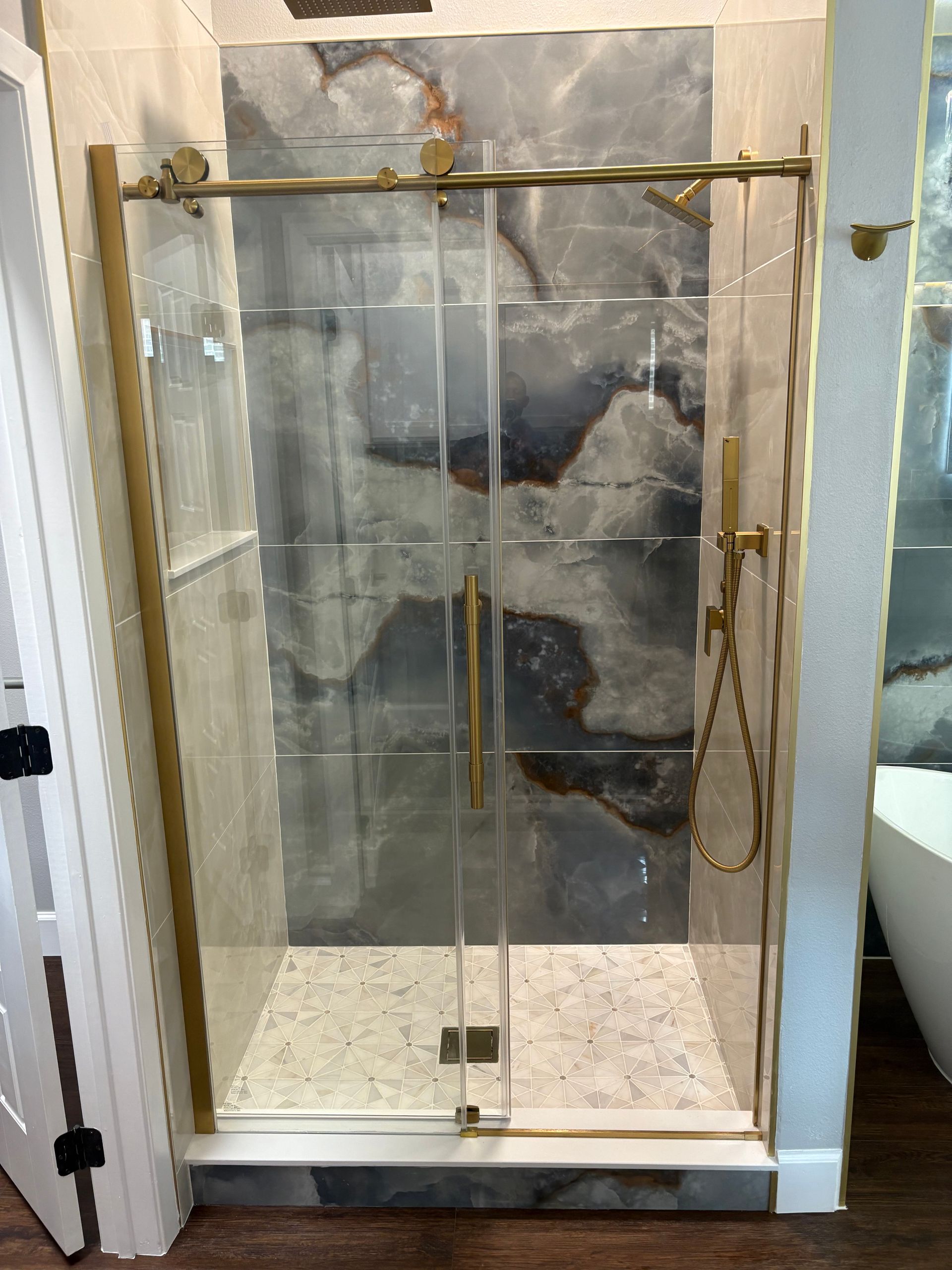 Gold-framed shower enclosure with sliding glass doors, marble-look tiled walls, and a pebble-stone shower floor.