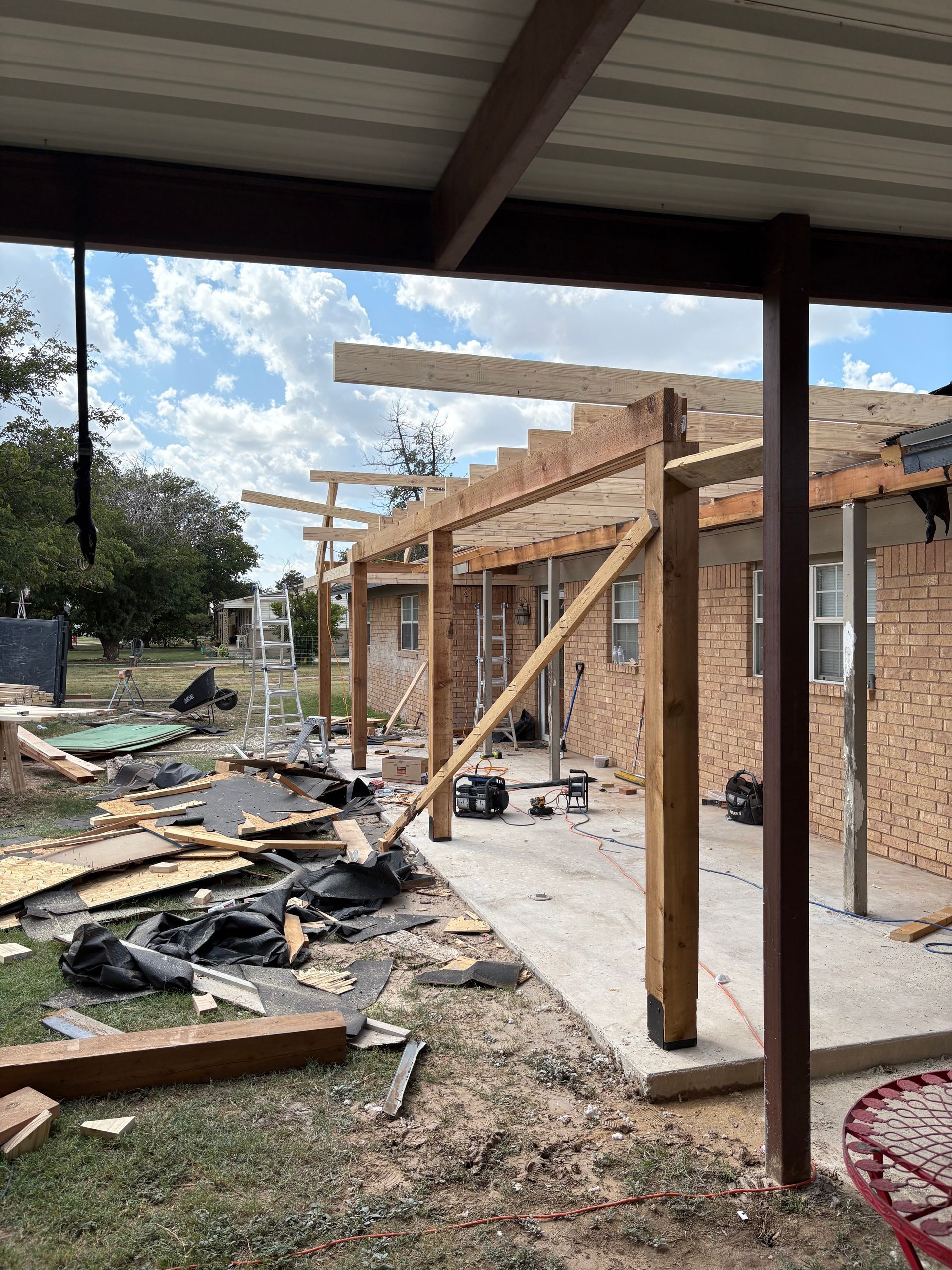 Patio under construction with exposed beams, lumber, tools. Brick house, blue sky visible.