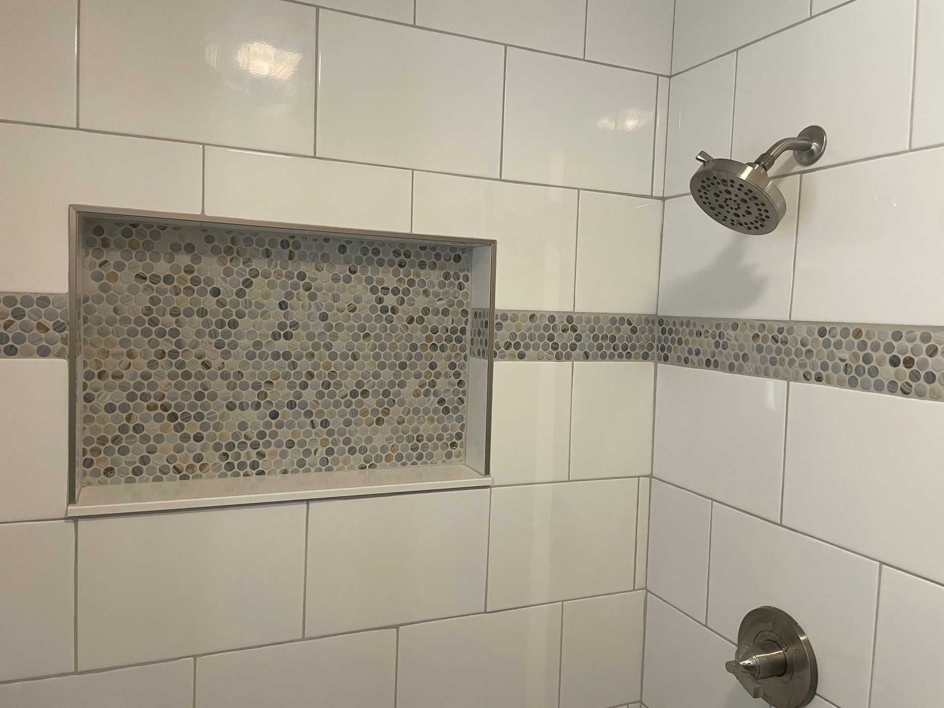 White tiled shower with a mosaic tile strip and built-in shelf. Silver shower head and faucet.