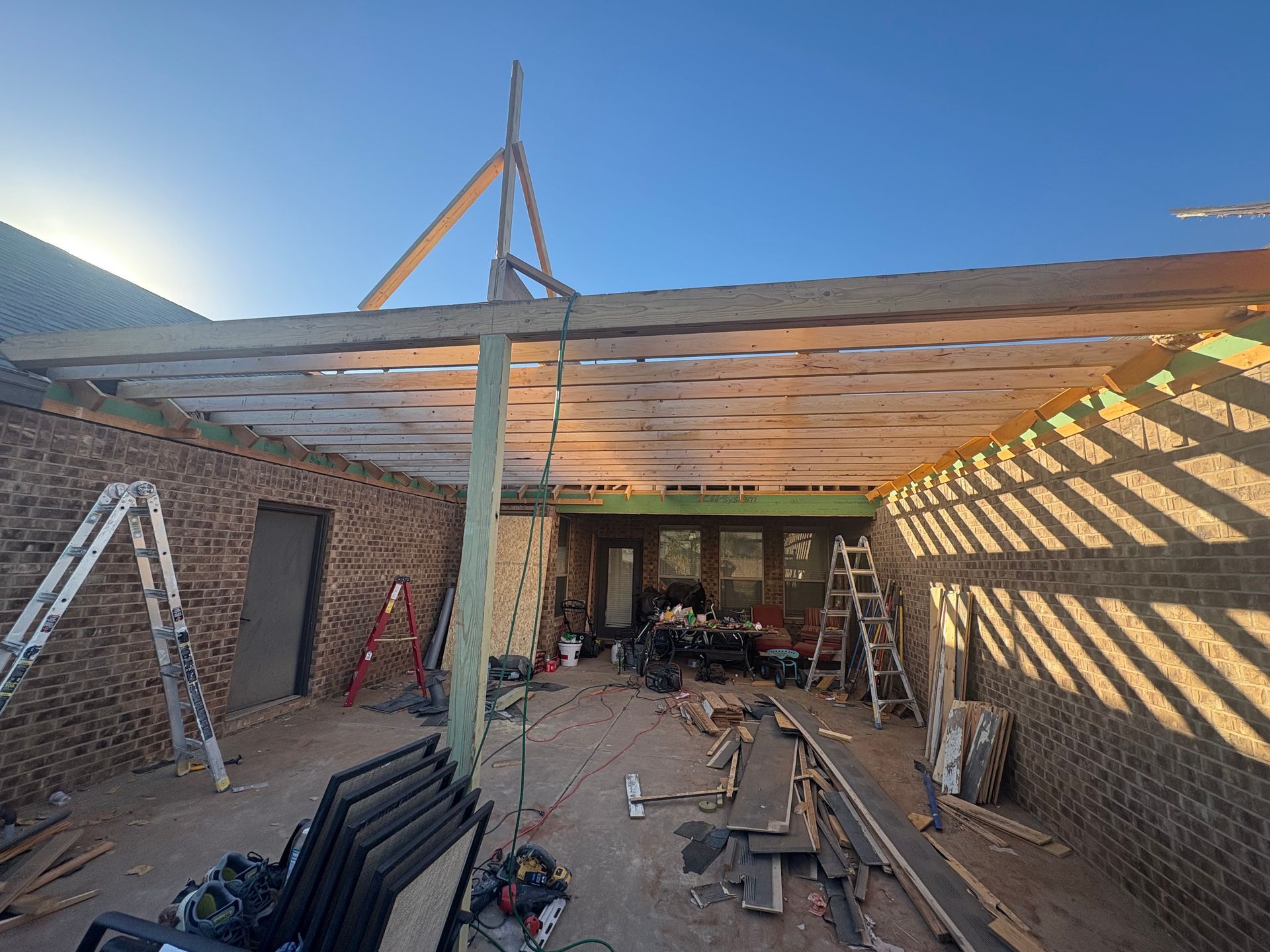 Construction site: Wooden beams and framing for a patio roof, with brick walls and equipment visible.