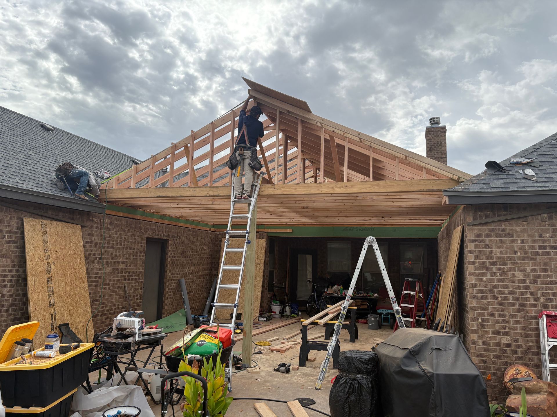 Construction workers building a wooden patio roof between two brick buildings under a cloudy sky.
