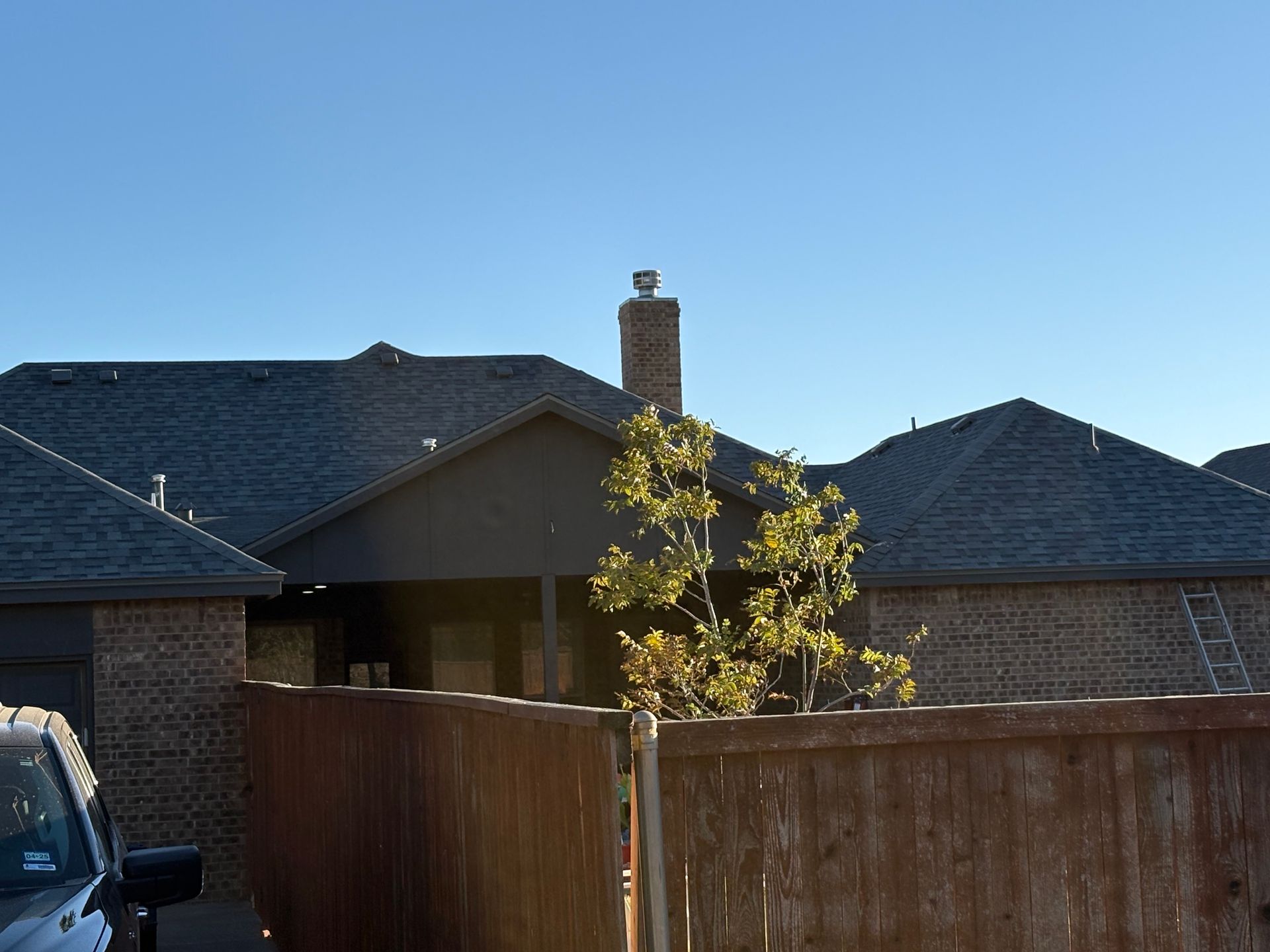 House with dark gray roof, brick exterior, wooden fence, tree, and blue sky.
