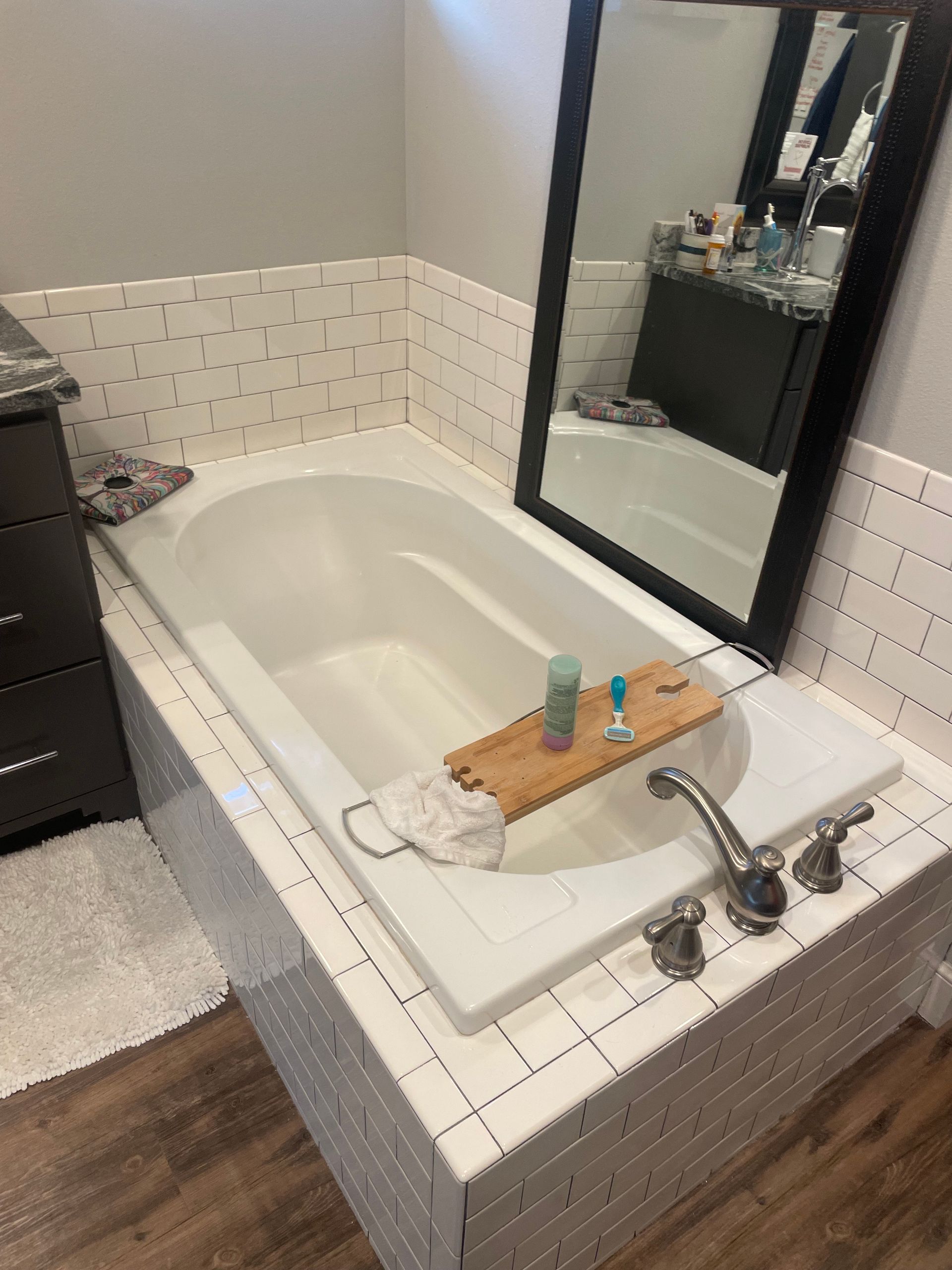 White bathtub with wood bath tray, set in tiled surround, with mirror reflecting a vanity.