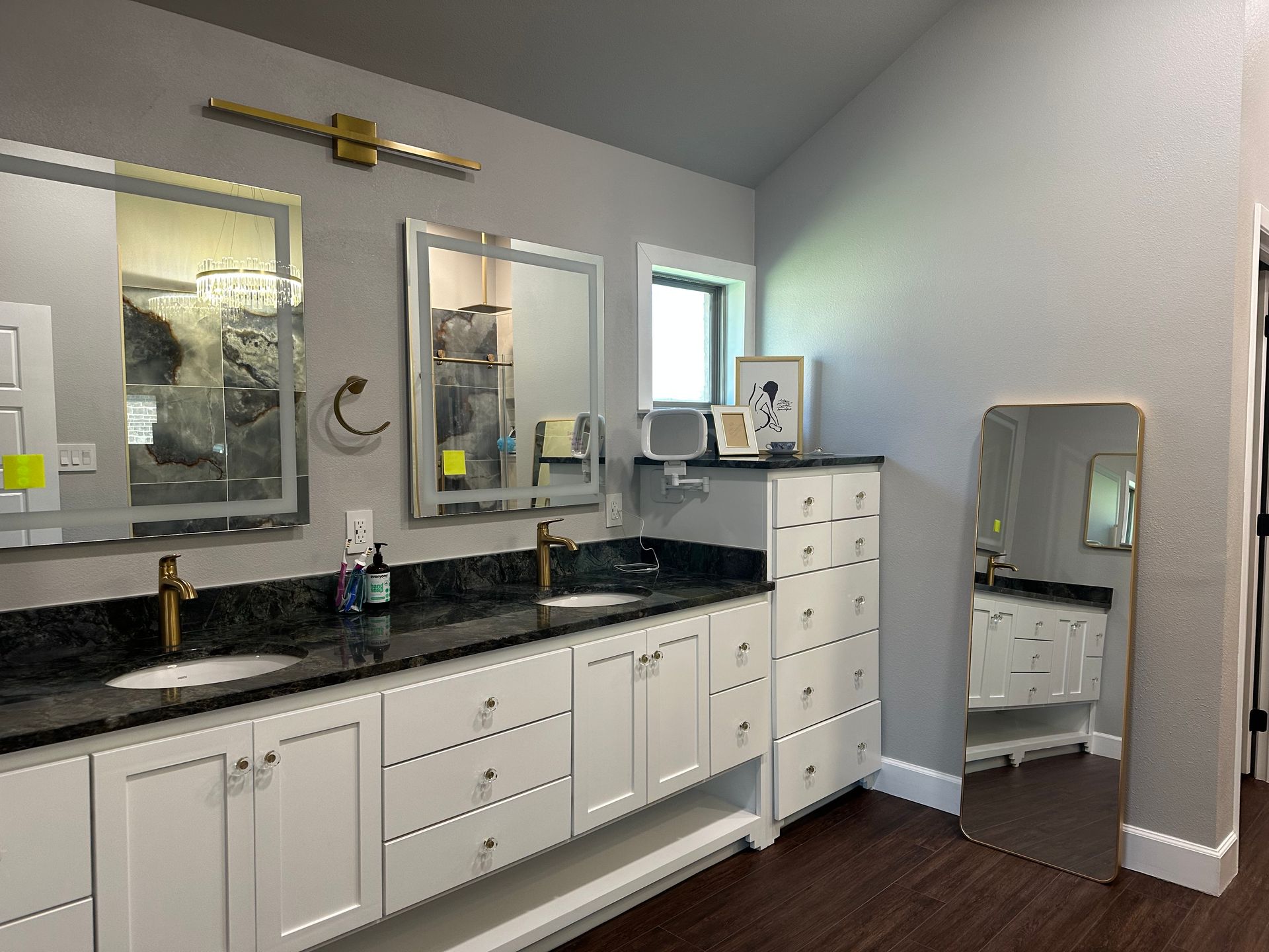 Bathroom with double sinks, white cabinets, gold fixtures, large mirrors, dark countertop, and standing mirror.