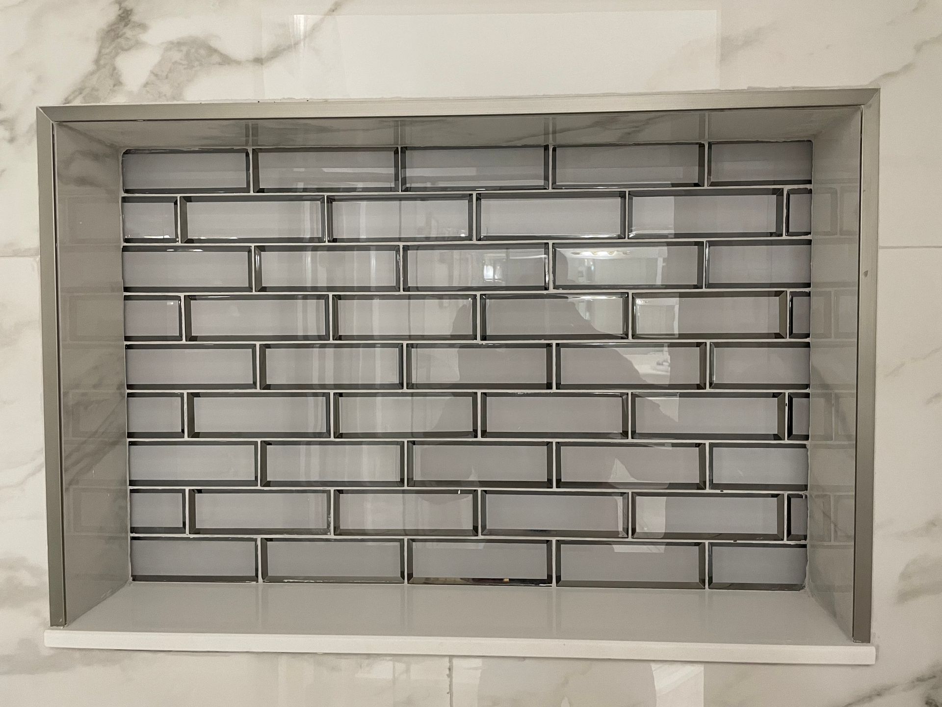 Recessed shower shelf with gray brick-pattern tile and chrome trim, set in a marble-look wall.