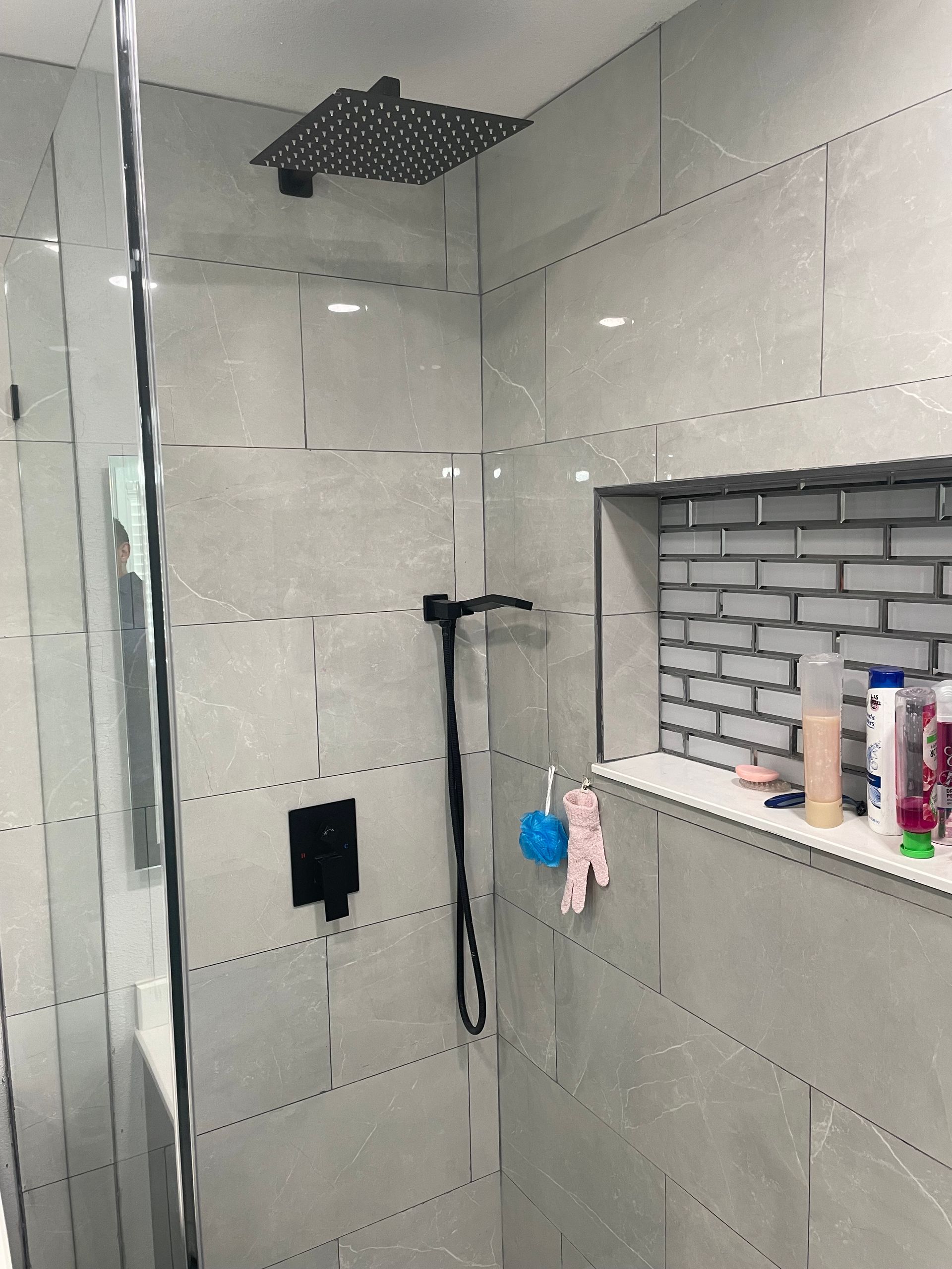 Modern shower with gray tiled walls, black fixtures, and a glass door. Built-in shelf with toiletries.