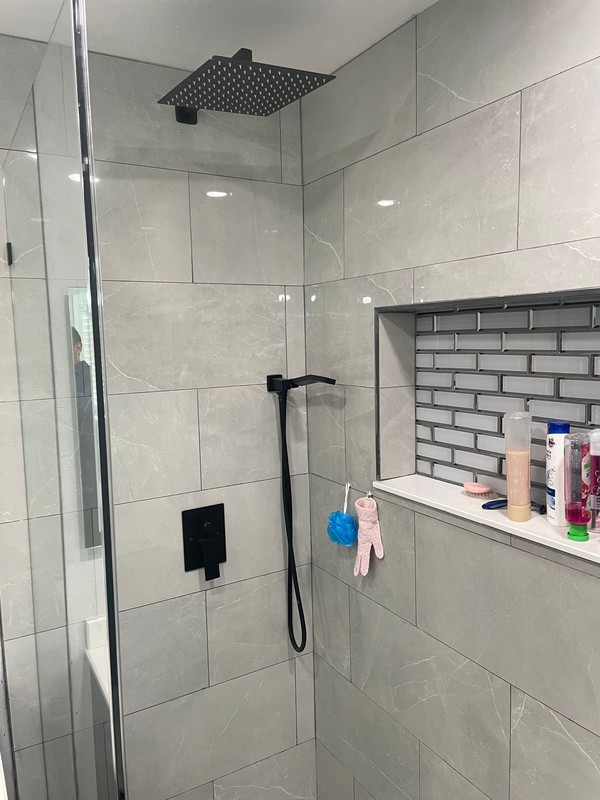 Modern shower with grey tiled walls, black fixtures, and a recessed shelf with toiletries.