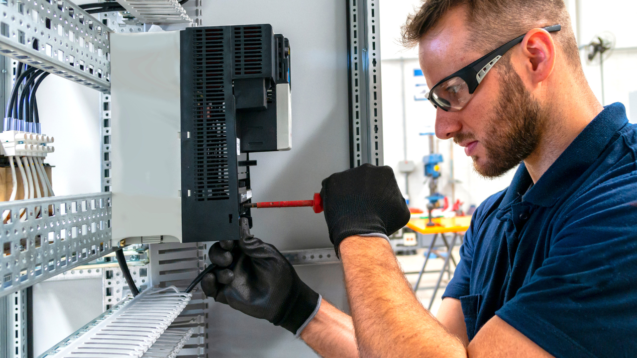 Man in safety glasses and gloves working on electrical panel with screwdriver.