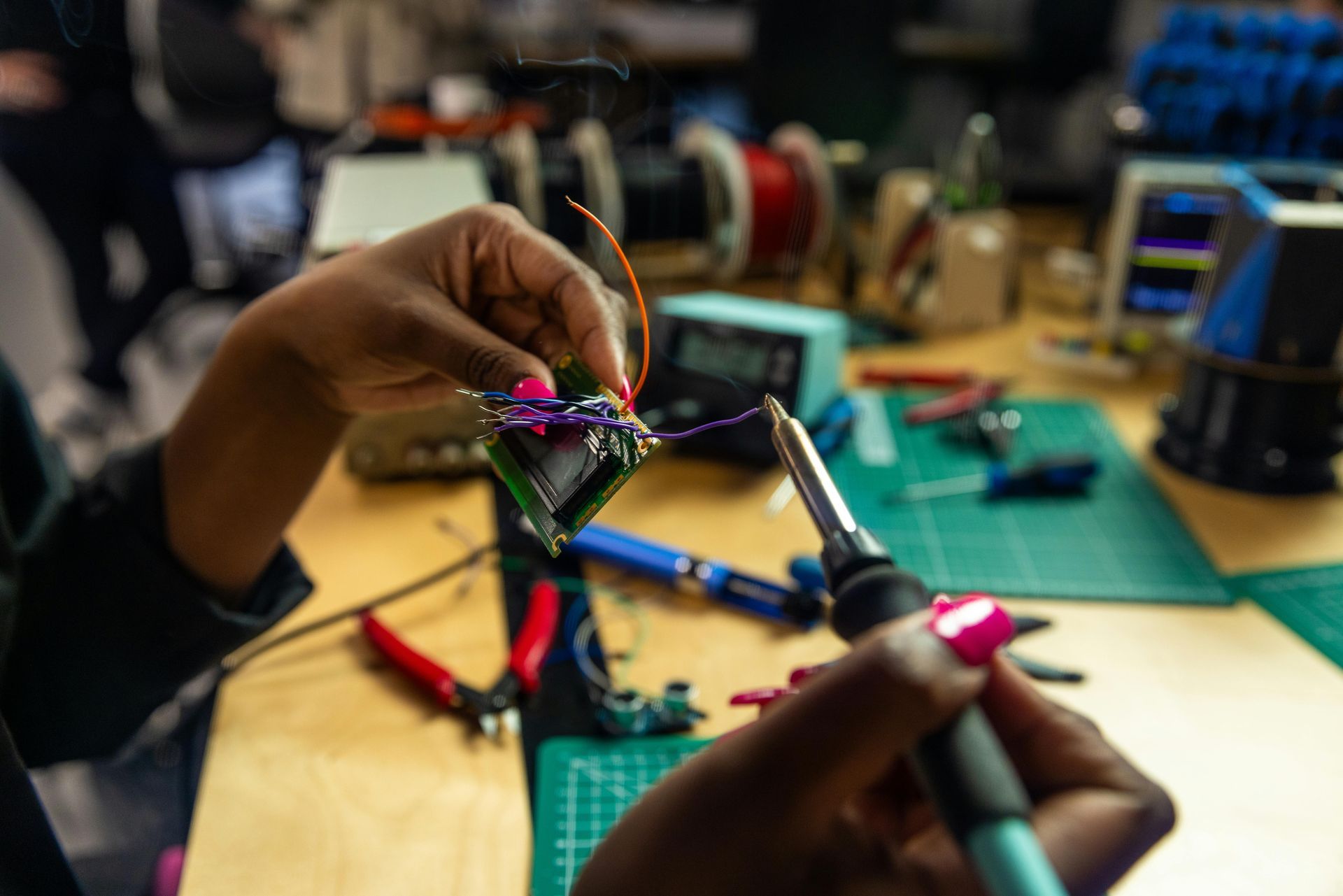 Hands soldering electronic components on a circuit board at a workbench, with tools and spools of wire.