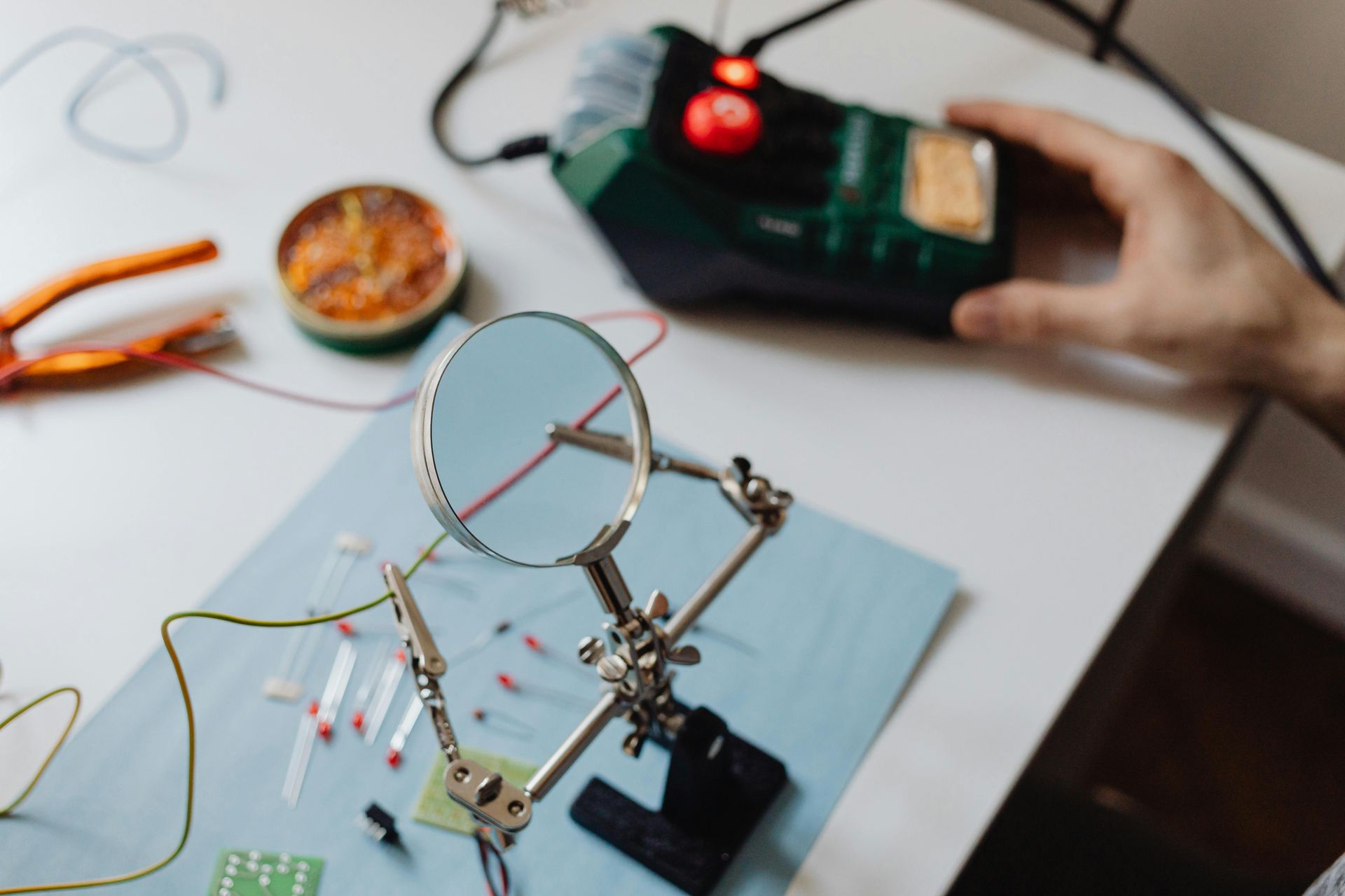 Person soldering electronics with magnifying glass, soldering iron, and components on a white surface.
