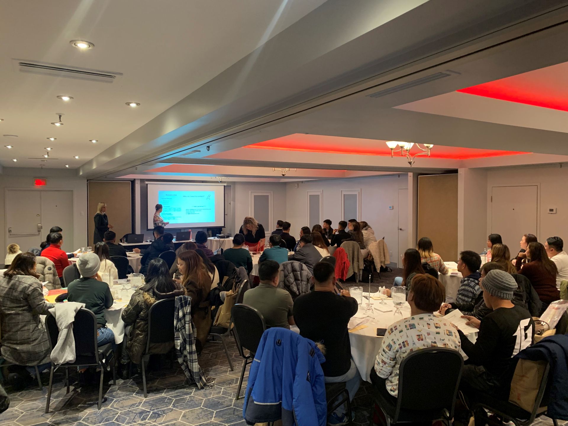 Un groupe de personnes sont assises à des tables dans une grande salle et regardent une présentation.