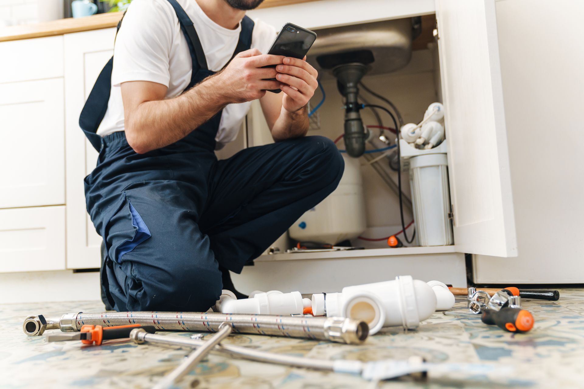 Plumber kneeling under a kitchen sink, checking a phone with pipes and tools nearby.