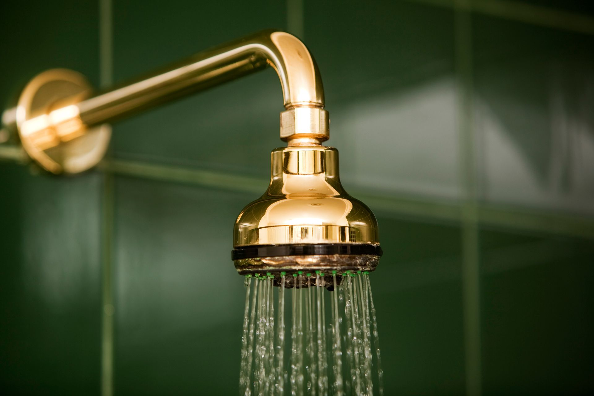 Gold showerhead with water spraying onto green tiled wall.
