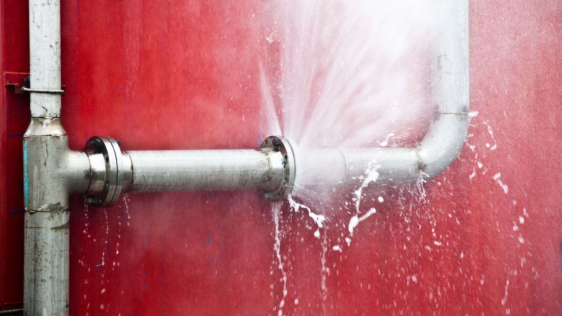 A metal pipe bursts, spraying water against a red wall.