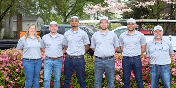 Group of seven people wearing matching gray shirts and hats, posing in front of a bush.