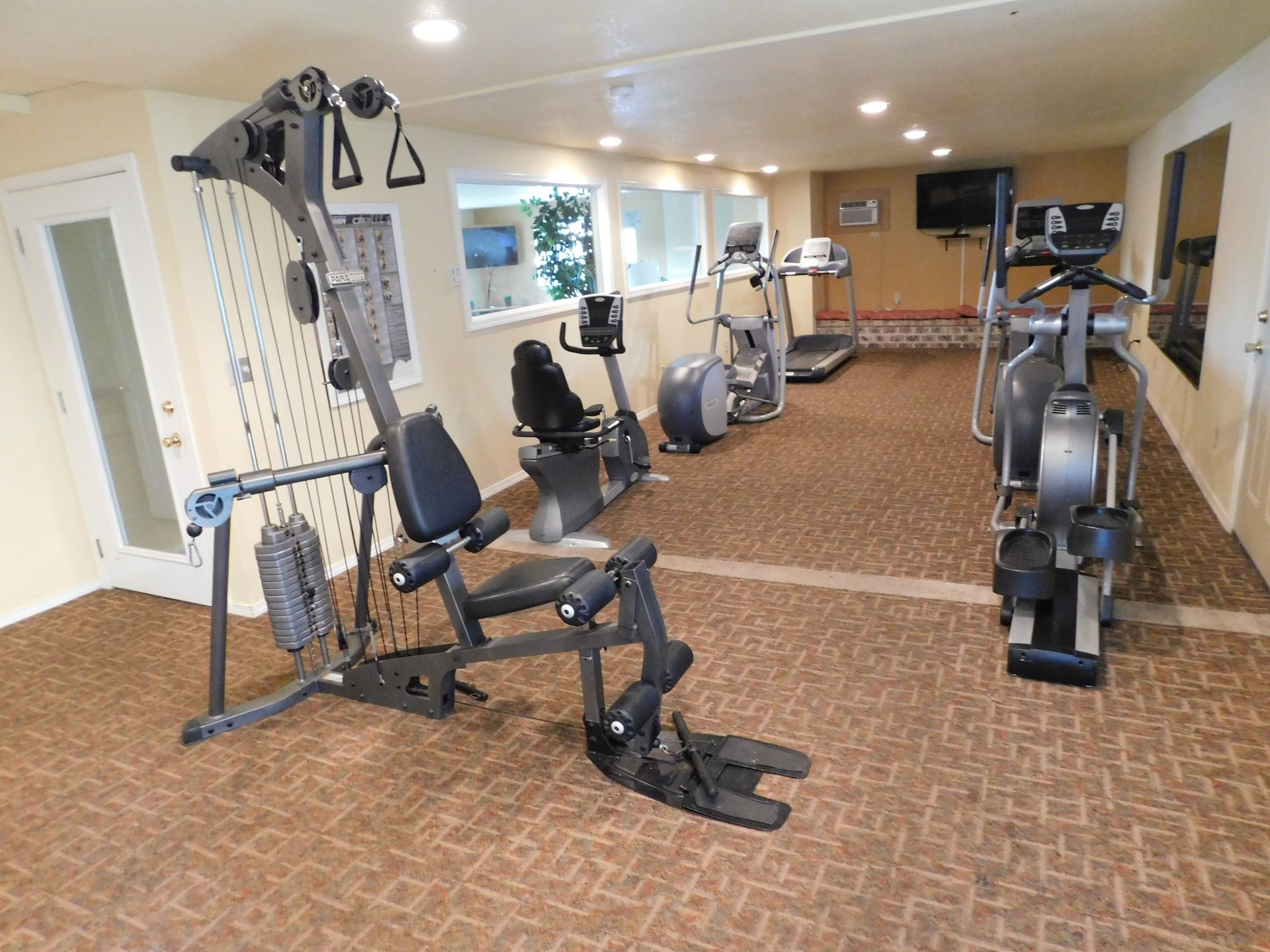 A fitness center with various exercise machines, including a weight machine, stationary bikes, and treadmills on a brown carpet.