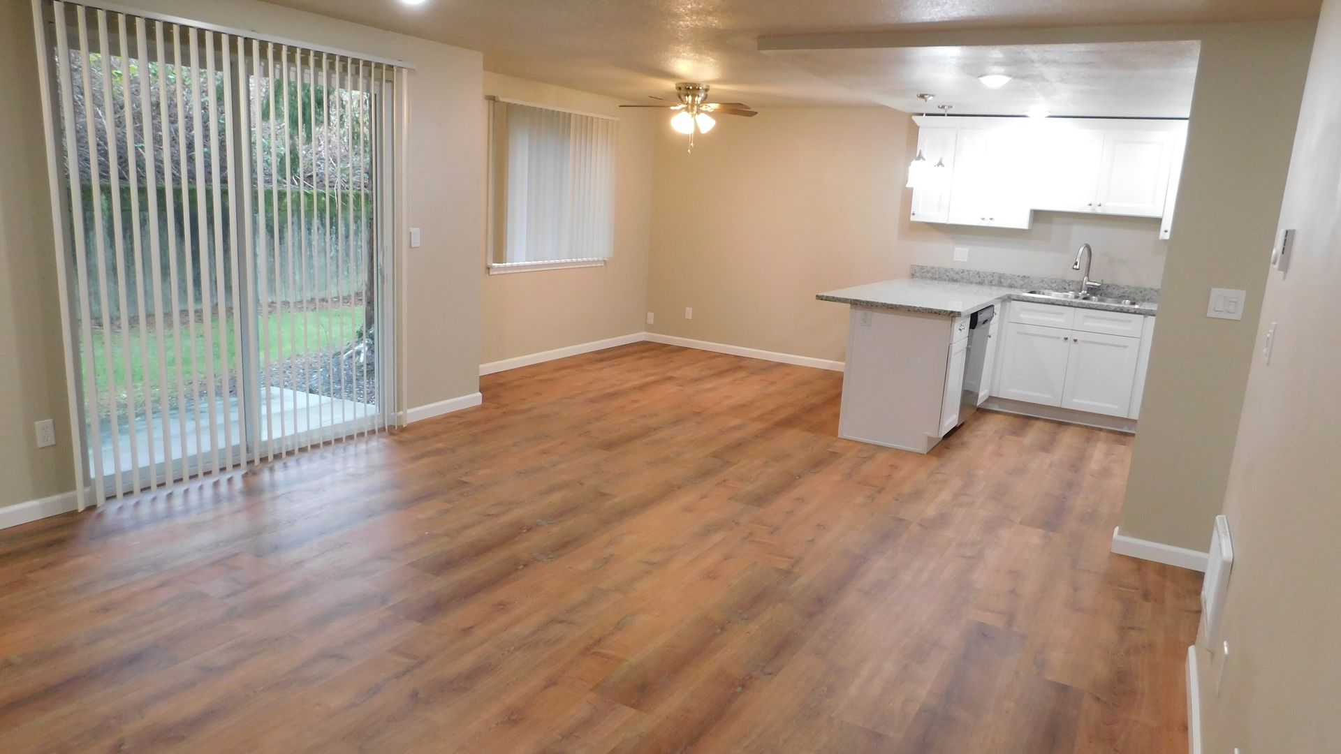 Empty living space with wood-look floors, sliding glass door, and partial kitchen. Beige walls and light.