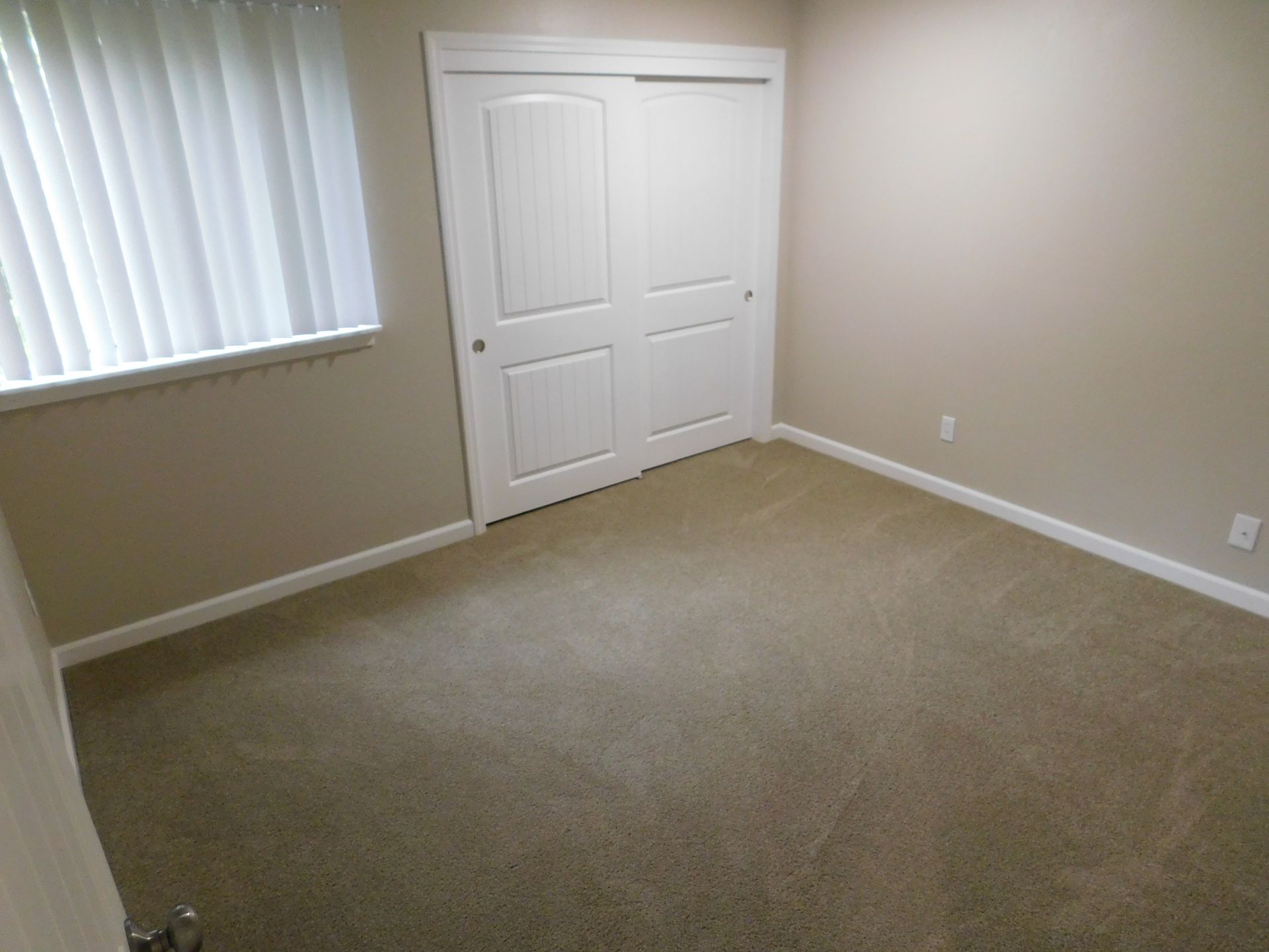 Empty bedroom with beige walls, closet, window with blinds, and carpet.