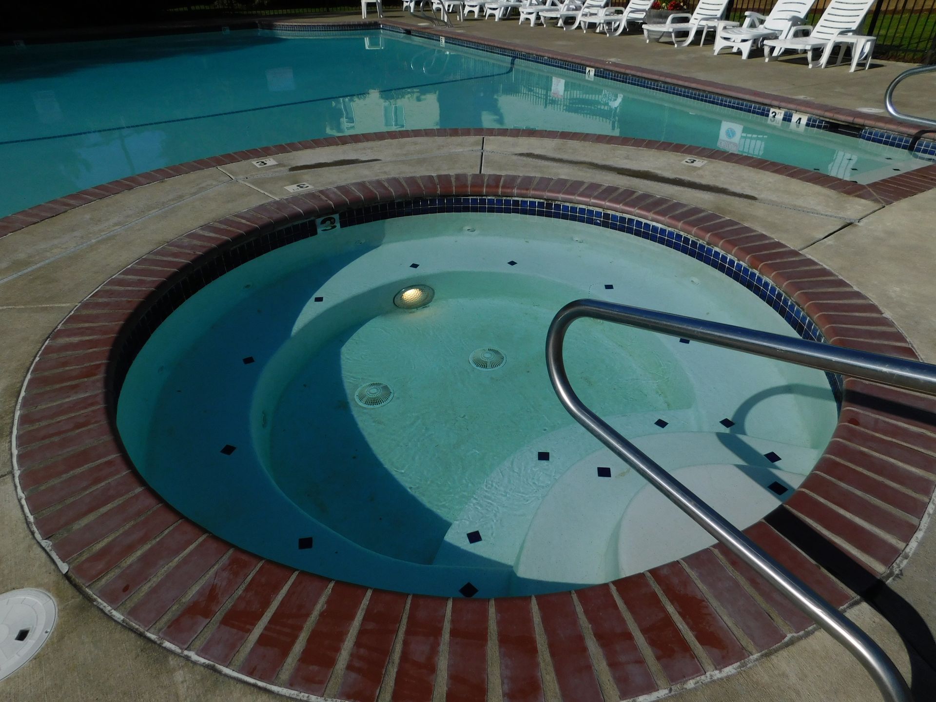 Hot tub next to a swimming pool, both outdoors. White chairs line the pool's edge.