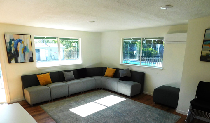 Living room with gray sectional sofa, rug, and colorful artwork. Windows let in sunlight.
