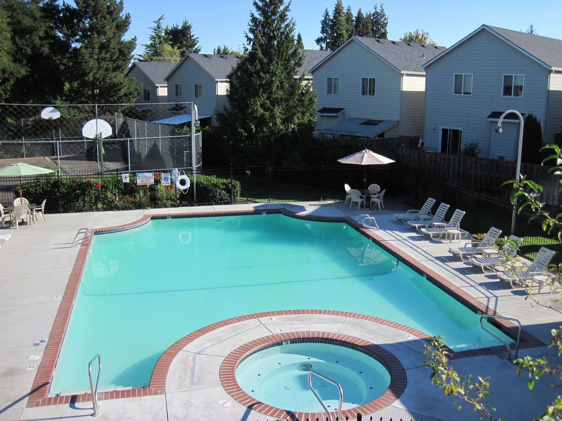 Swimming pool and hot tub with lounge chairs, next to a community of gray houses.
