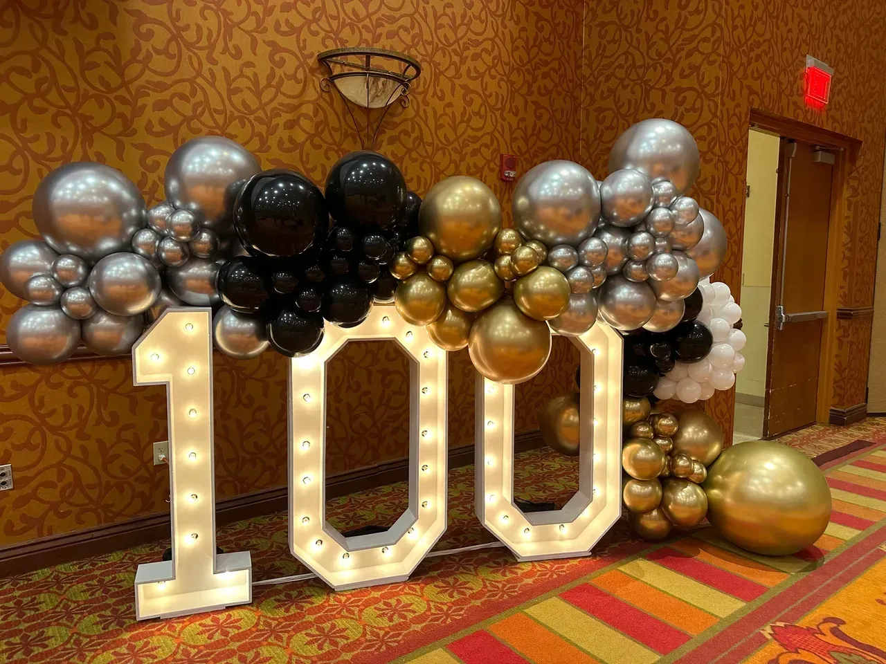 A large number 100 is surrounded by balloons in a room.